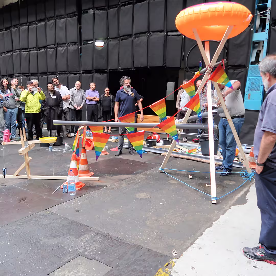 A group of delegates at the Dyalog '17 event in Elsinore, Denmark, watch as participants work on a large, makeshift contraption decorated with rainbow bunting. The setup includes wooden structures, pipes, traffic cones, and an inflatable orange ring suspended at the top. The scene takes place indoors in a large industrial-style space with black padded walls. Some delegates observe from a distance, while others actively engage with the structure.