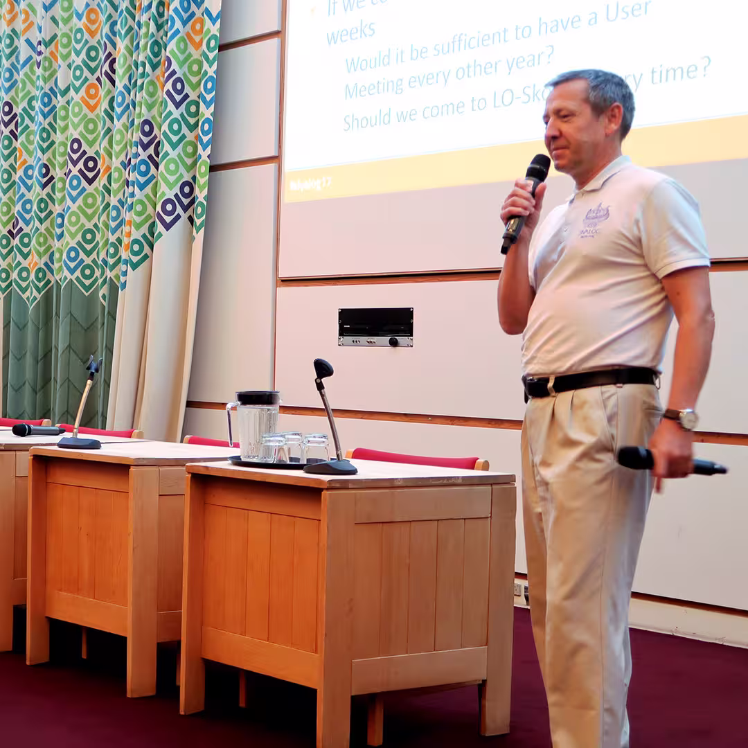 Morten Kromberg, wearing a white Dyalog-branded polo shirt and beige trousers, speaks into a handheld microphone during the Dyalog '17 event at the Konventum Convention Centre in Elsinore, Denmark. He holds a second microphone in his left hand. Behind him, a projector screen displays discussion points about the frequency and location of future user meetings. To the left, a table with microphones, a water jug, and glasses sits in front of vibrant, patterned curtains.