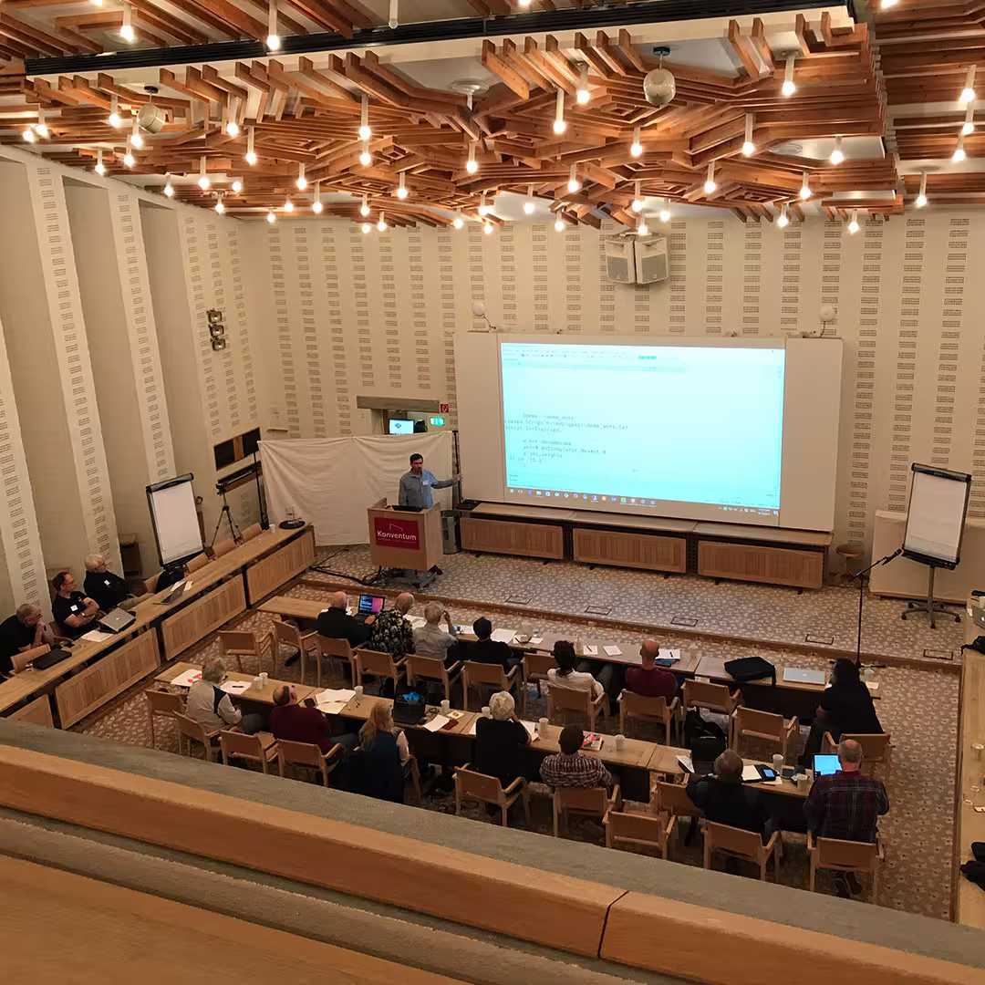 A presentation at the Dyalog '17 event held at the Konventum Convention Centre in Elsinore, Denmark. A speaker stands at a red Konventum-branded podium, gesturing toward a large projection screen displaying APL code. The room has modern wooden ceiling panels with hanging light bulbs. Several rows of delegates sit at tables, some taking notes on laptops. Two flipcharts stand on either side of the room.