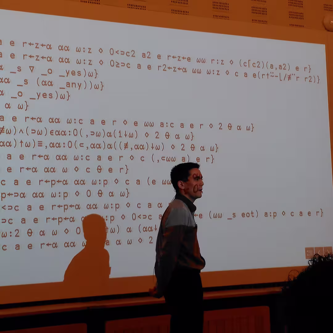 Aaron Hsu presents at the Dyalog '17 event in Elsinore, Denmark, standing in front of a large projection screen filled with APL code. The projection casts the code onto his face and clothing, creating a striking visual effect. He stands with his hands behind his back, attentively observing the audience. The light-colored wall behind the screen features subtle perforated paneling typical of the Konventum Convention Centre.