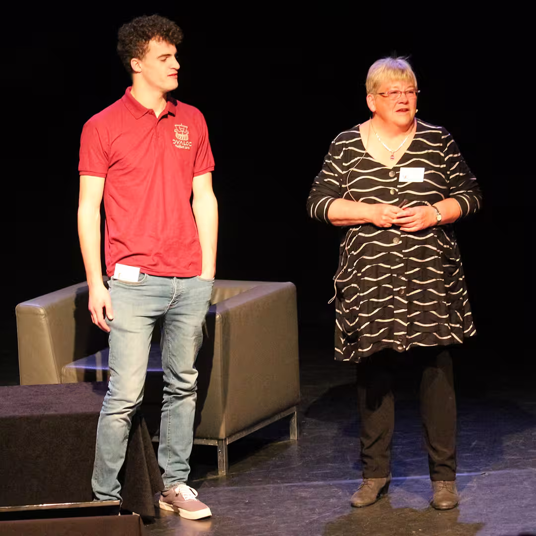Gitte Christensen speaking on stage during the Dyalog '18 event in Belfast, Northern Ireland. She is wearing a black and white patterned dress, black trousers, and glasses. Next to her stands a young man wearing a burgundy Dyalog-branded polo shirt and jeans. Both are standing near two grey armchairs, with a dark background behind them.