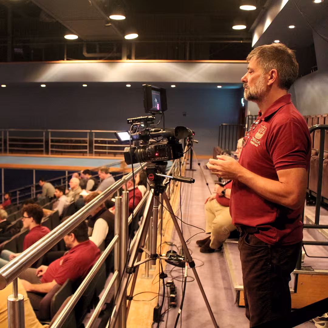 Bjørn Christensen operating a video camera during the Dyalog '18 event in Belfast, Northern Ireland. He is wearing a burgundy Dyalog-branded polo shirt and standing behind a professional video camera mounted on a tripod, recording the stage. The background shows audience members seated in the auditorium.