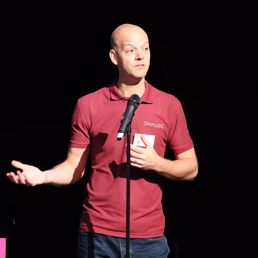 Jay Foad speaking into a microphone during the Dyalog '18 event in Belfast, Northern Ireland. He is wearing a burgundy Dyalog-branded polo shirt and gesturing with one hand. The background is dark.