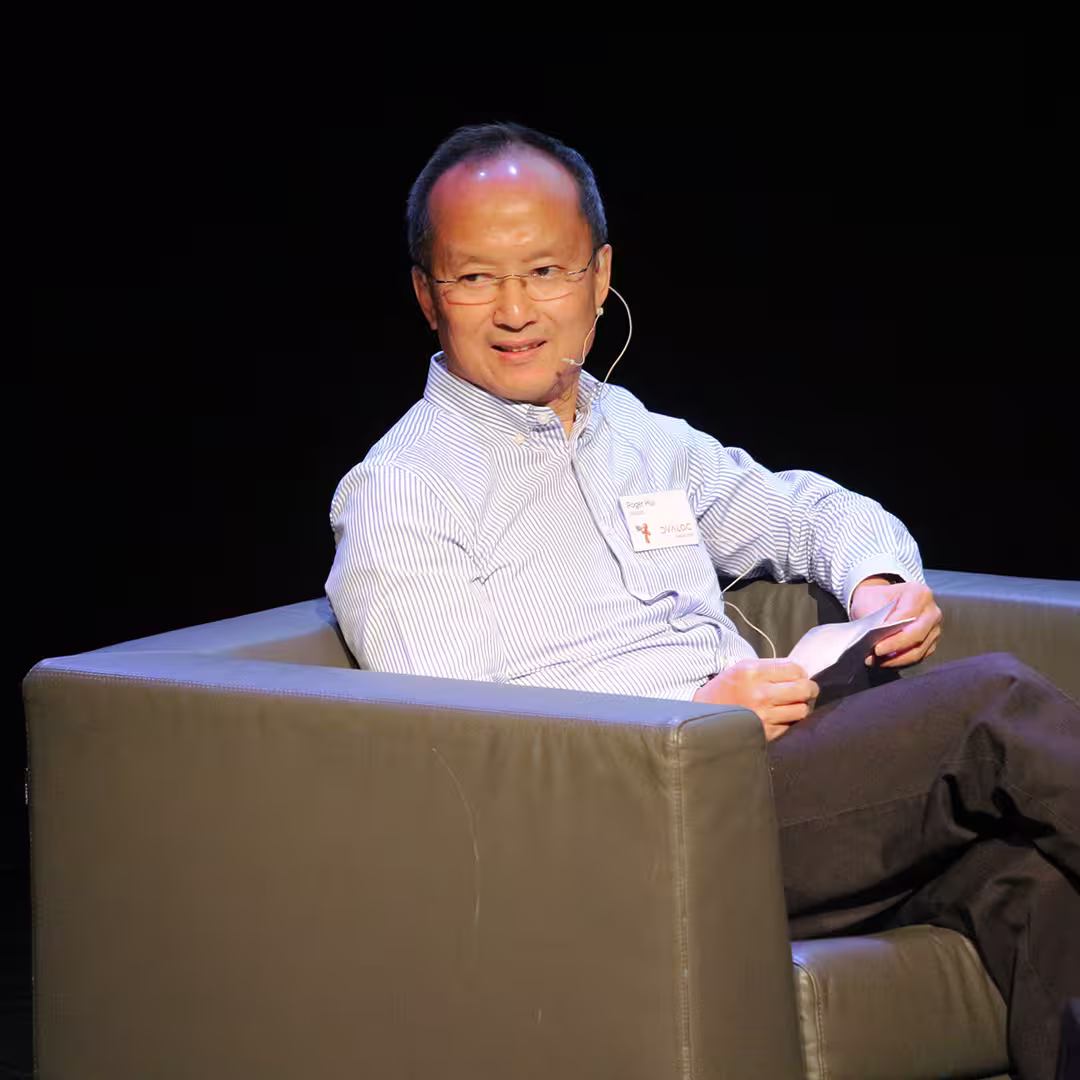 Roger Hui seated in a grey armchair during the Dyalog '18 event in Belfast, Northern Ireland. He is wearing a light blue striped shirt, dark trousers, and a headset microphone. He holds a piece of paper and appears to be speaking or listening, with a dark background behind him.