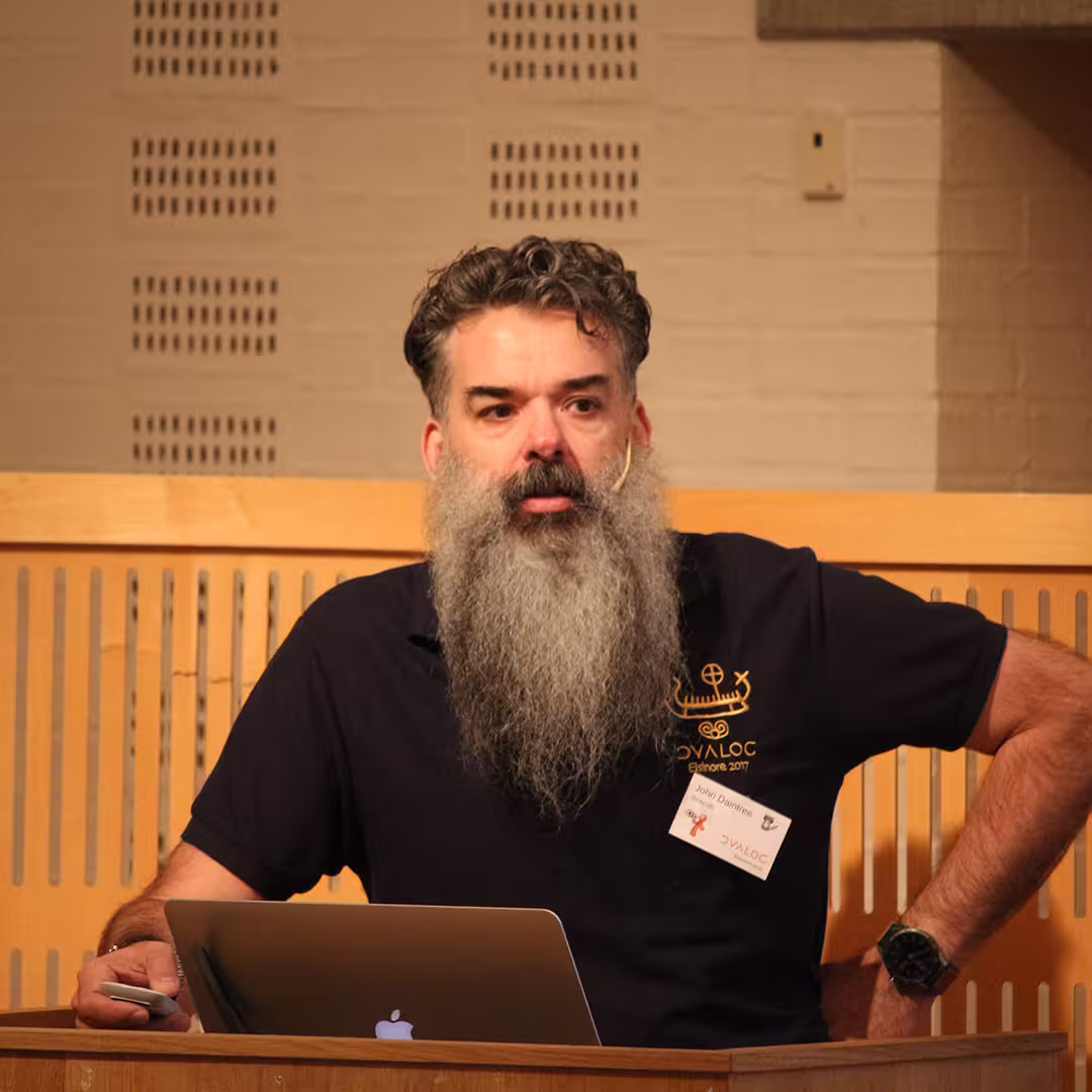 John Daintree presenting at Dyalog '19, held at the Konventum Convention Centre in Elsinore, Denmark. He stands at the lectern, wearing a Dyalog-branded polo shirt and a name badge, speaking into a headset microphone with a laptop open in front of him.