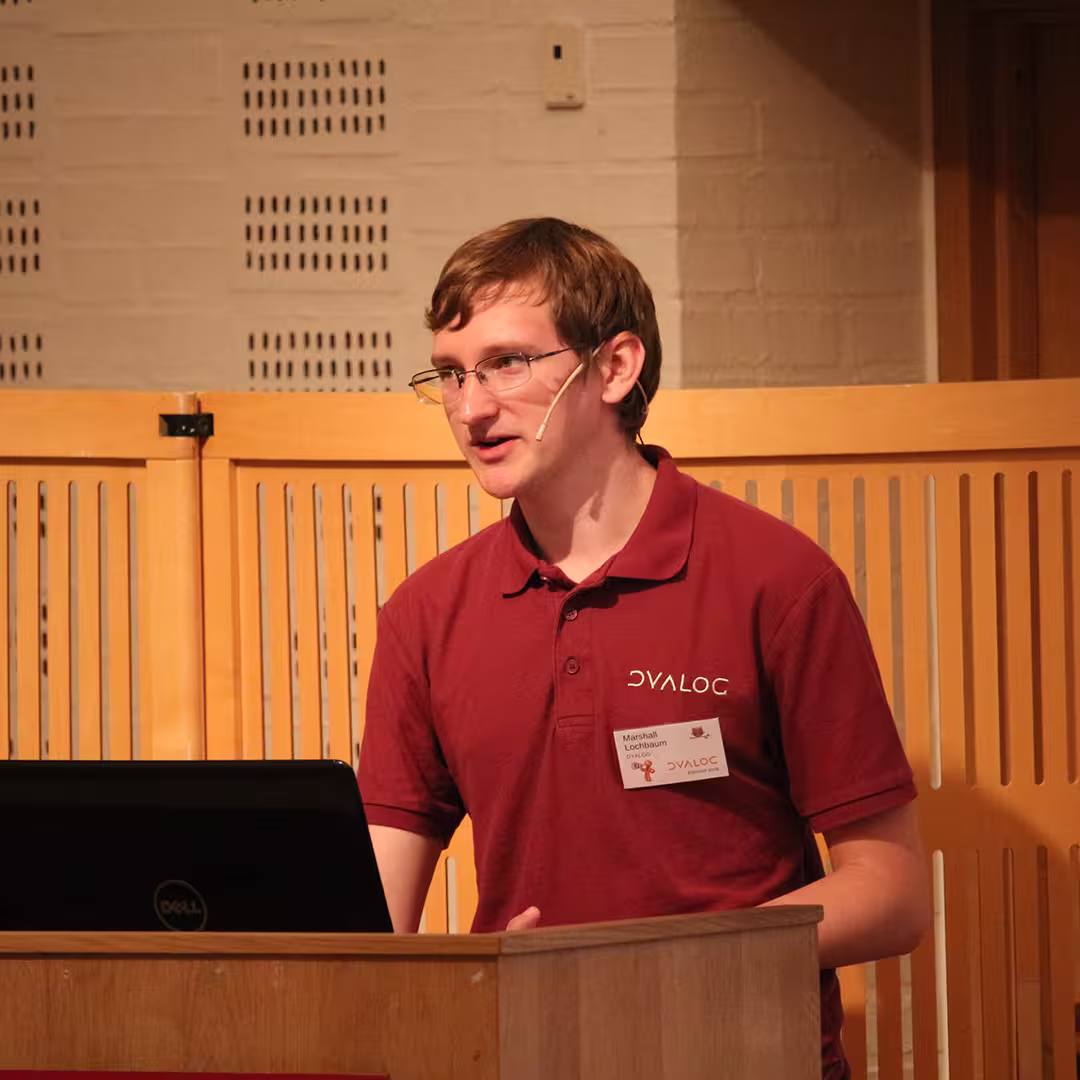 Marshall Lochbaum presenting at Dyalog '19, held at the Konventum Convention Centre in Elsinore, Denmark. He stands at the lectern, wearing a Dyalog-branded polo shirt and a name badge, speaking into a headset microphone with a laptop open in front of him.