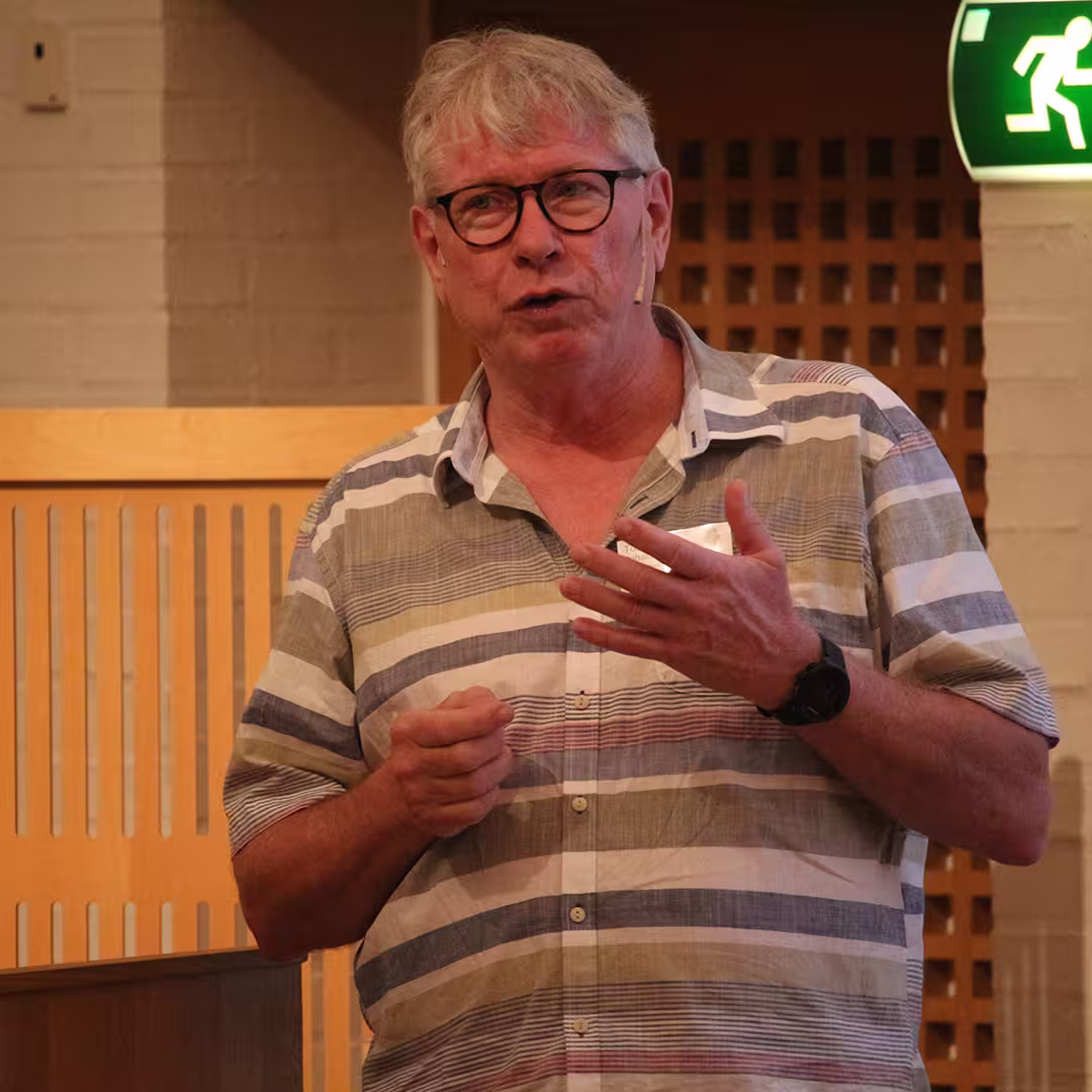 A delegate presenting at Dyalog '19, held at the Konventum Convention Centre in Elsinore, Denmark. He is speaking into a headset microphone while gesturing with his hands, wearing a striped shirt and glasses.
