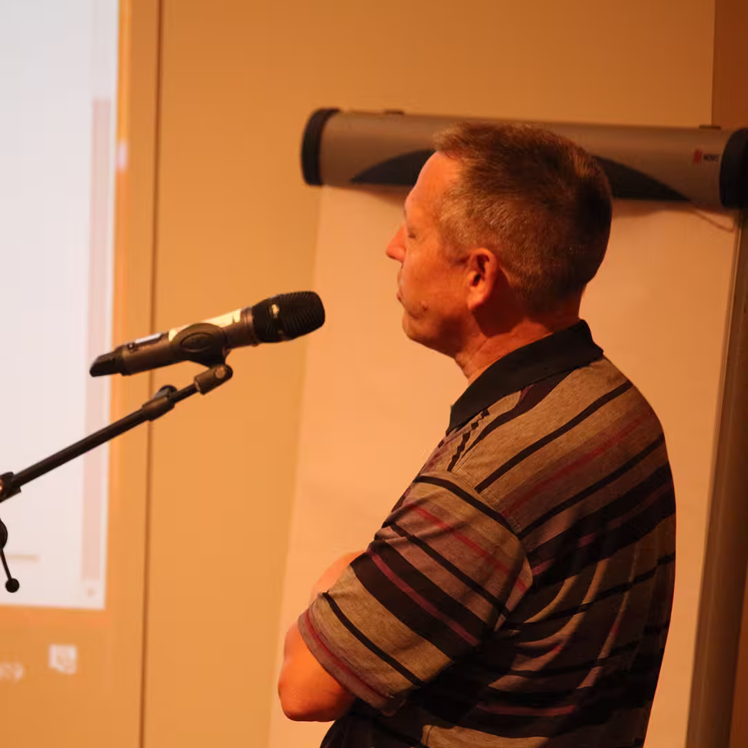 Morten Kromberg presenting at Dyalog '19, held at the Konventum Convention Centre in Elsinore, Denmark. He stands at a microphone, speaking while looking at the projected presentation screen in front of a flip chart.