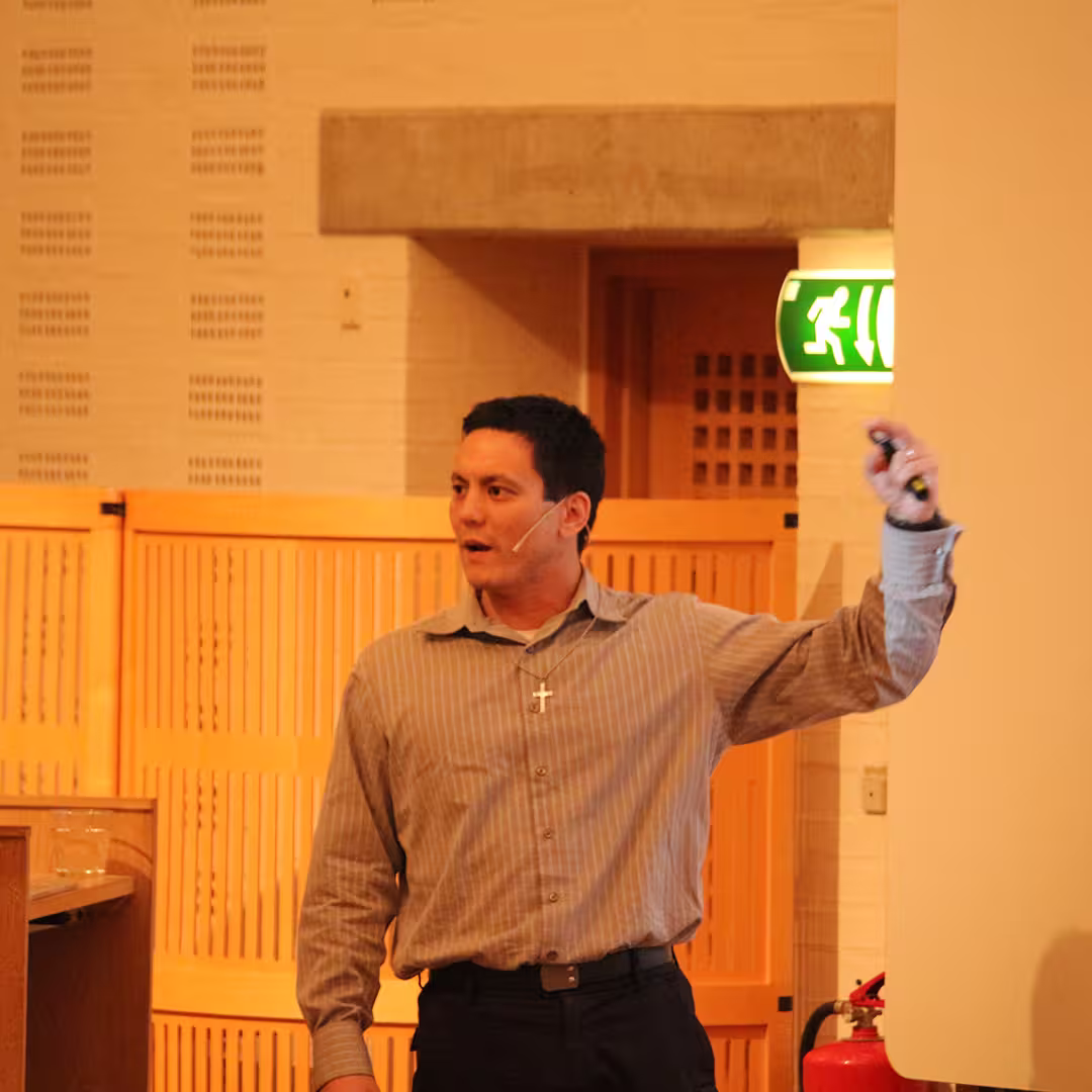 Aaron Hsu presenting at Dyalog '19, held at the Konventum Convention Centre in Elsinore, Denmark. He is speaking into a headset microphone, gesturing with one hand while holding a presentation remote in the other.