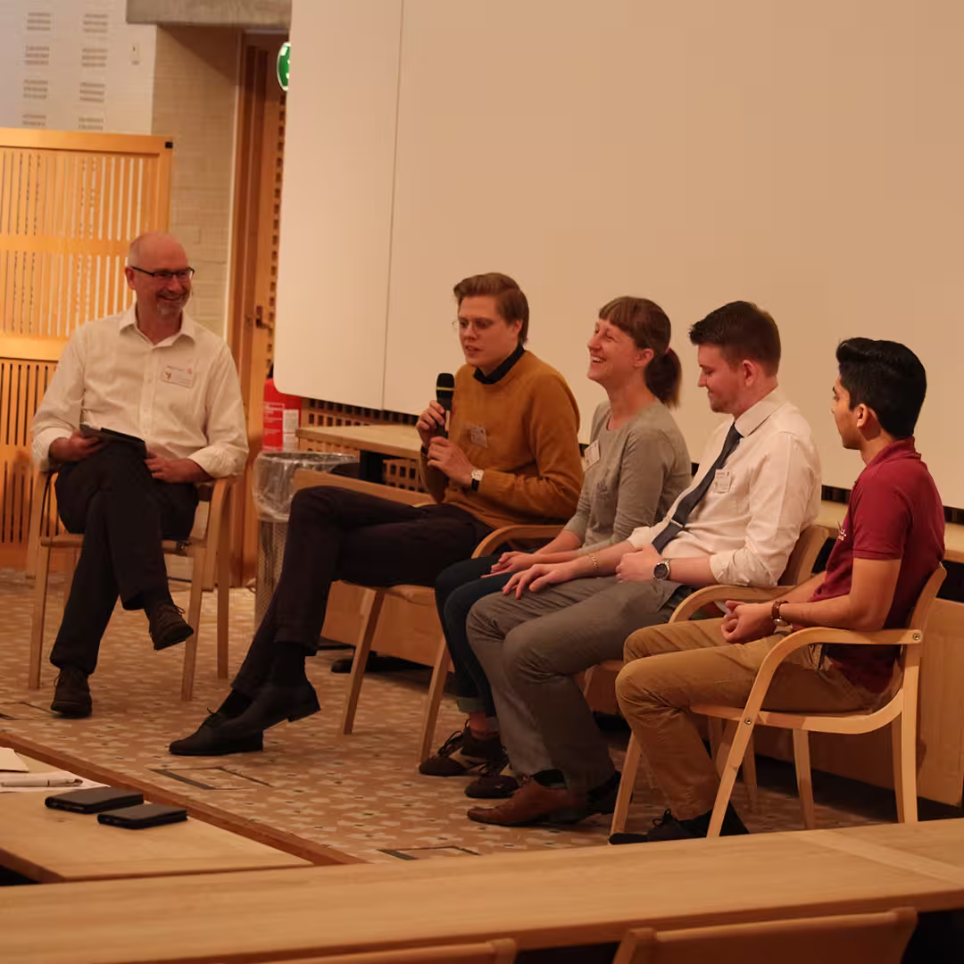 Stephen Taylor (far left) moderating a panel discussion at Dyalog '19, held at the Konventum Convention Centre in Elsinore, Denmark. Panelists include Josh David (far right), James Heslip (second from right), and two other delegates seated between them. One panelist speaks into a microphone while the others listen and smile.