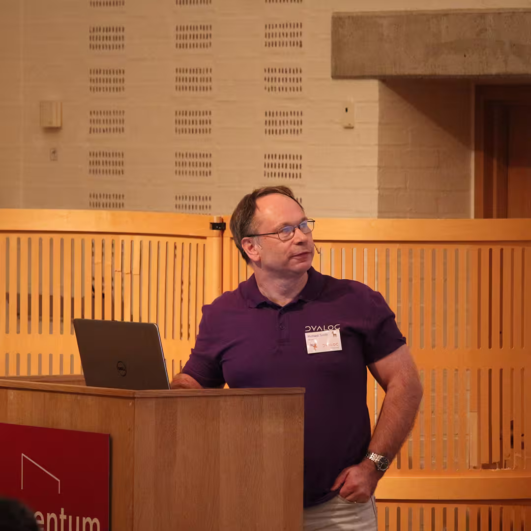 Richard Smith presenting at Dyalog '19, held at the Konventum Convention Centre in Elsinore, Denmark. He stands at the lectern, wearing a purple Dyalog-branded polo shirt and a name badge, with a laptop open in front of him.