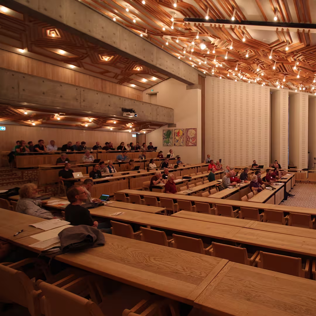 Audience members seated in the conference hall at Dyalog '19, held at the Konventum Convention Centre in Elsinore, Denmark. The room features wooden benches, a distinctive wooden ceiling design with hanging lights, and a concrete balcony area where more delegates are seated.