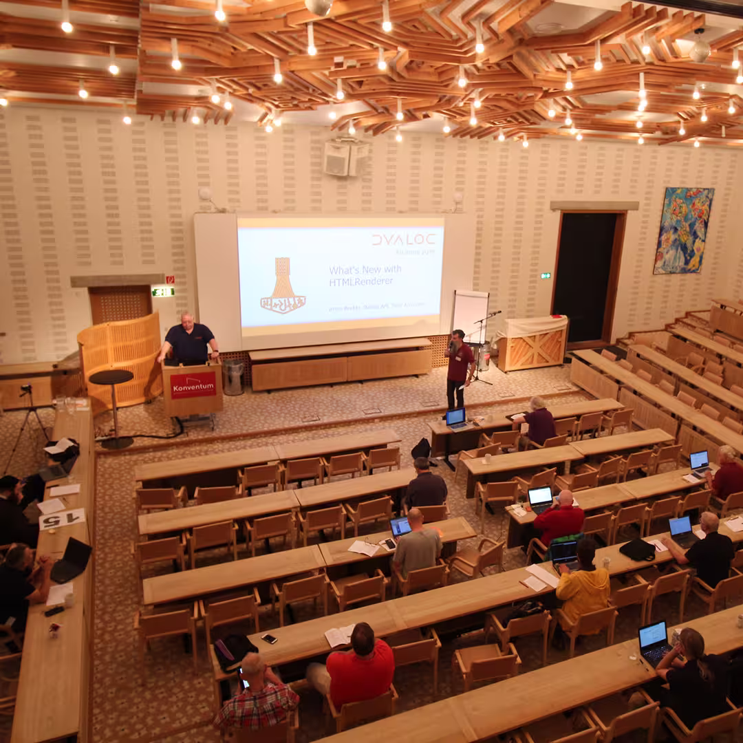 A presentation at Dyalog '19, held at the Konventum Convention Centre in Elsinore, Denmark. Brian Becker stands at the lectern while another delegate presents on stage. The slide projected behind them reads "What's New with HTMLRenderer." Audience members are seated with laptops in the conference hall, which features a distinctive wooden ceiling design and bright lighting.