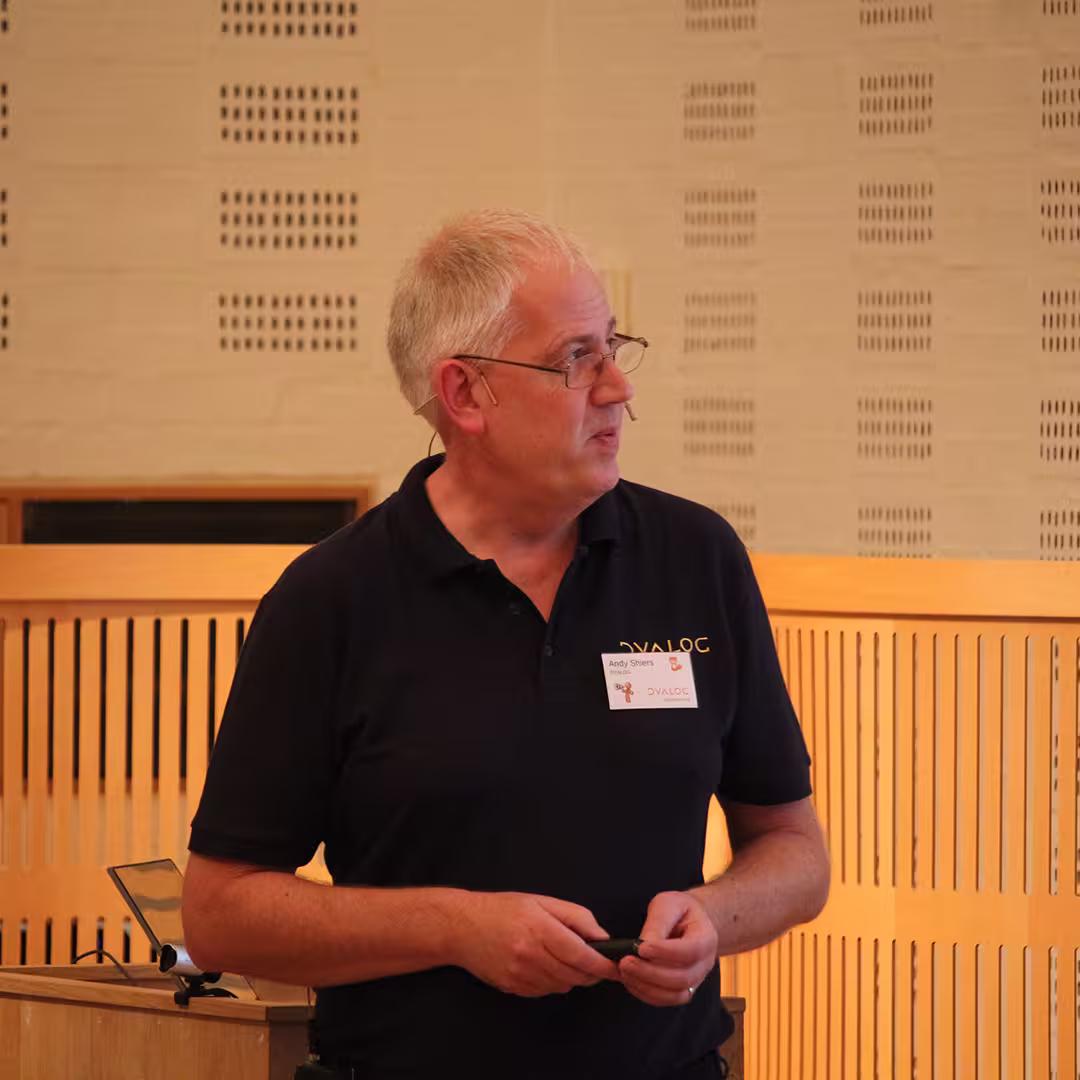 Andy Shires presenting at Dyalog '19, held at the Konventum Convention Centre in Elsinore, Denmark. He is standing at the lectern, wearing a Dyalog-branded polo shirt and a name badge, while holding a presentation remote.