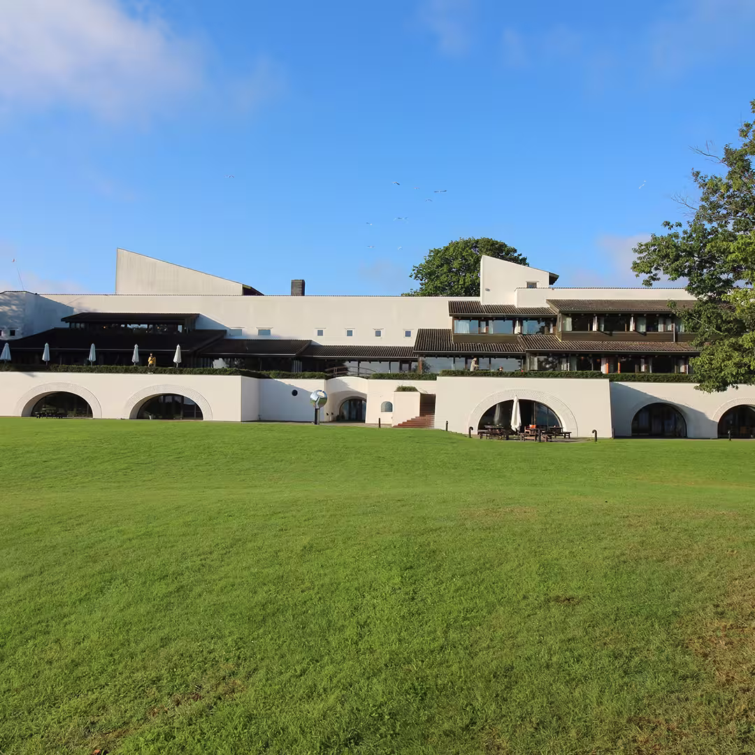 Exterior view of the Konventum Convention Centre in Elsinore, Denmark, the venue for Dyalog '19. The modern white building features arched windows, terraces with sun umbrellas, and a large green lawn in the foreground under a clear blue sky.