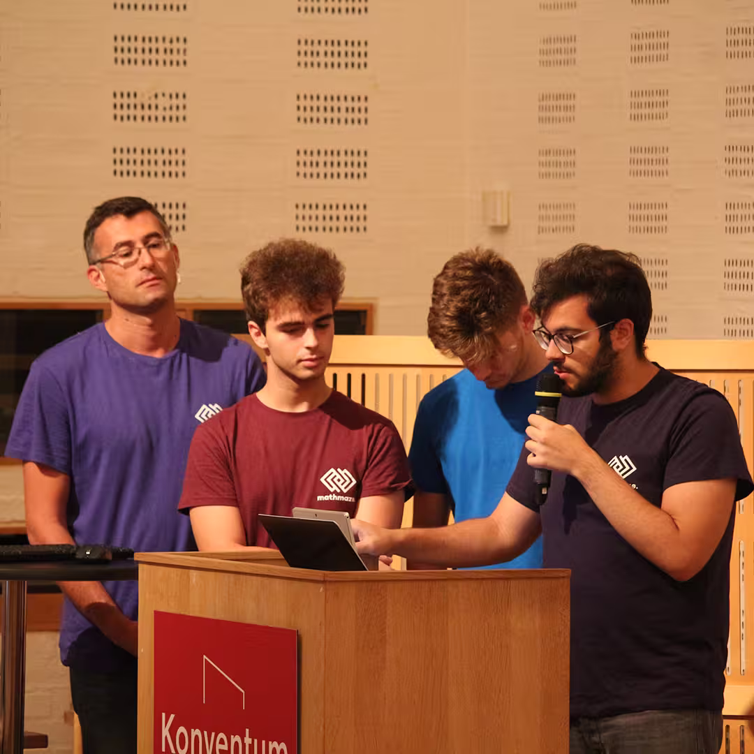 Four delegates presenting at Dyalog '19, held at the Konventum Convention Centre in Elsinore, Denmark. One delegate speaks into a microphone while using a tablet at the lectern, which displays the Konventum logo. The other three stand behind him, attentively observing the presentation.