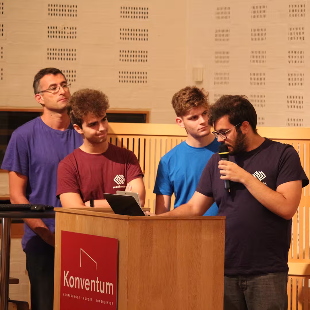 A group of four delegates presenting at Dyalog '19, held at the Konventum Convention Centre in Elsinore, Denmark. One delegate speaks into a microphone while using a tablet at the lectern, which features the Konventum logo. The other three stand attentively behind him.