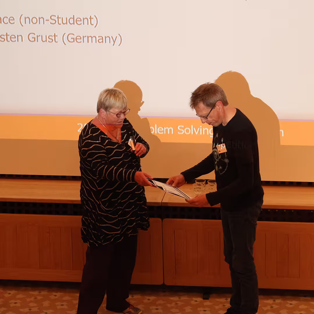 Gitte Christensen presenting a certificate to a delegate on stage at Dyalog '19, held at the Konventum Convention Centre in Elsinore, Denmark. A projection screen behind them displays competition results.