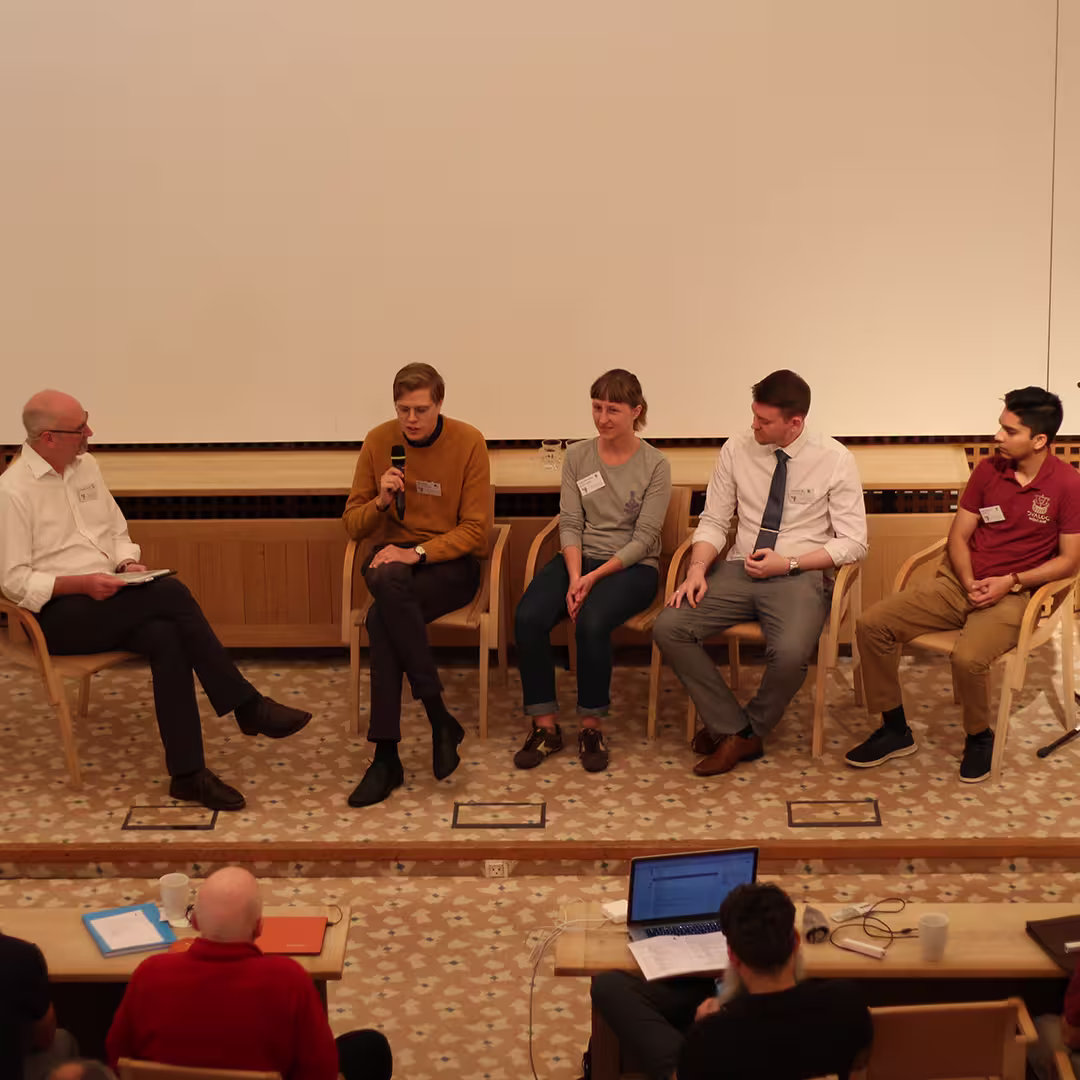 Stephen Taylor (far left) moderating a panel discussion at Dyalog '19, held at the Konventum Convention Centre in Elsinore, Denmark. Panelists include Josh David (far right), James Heslip (second from right), and two other delegates seated between them. Audience members and a laptop are visible in the foreground.
