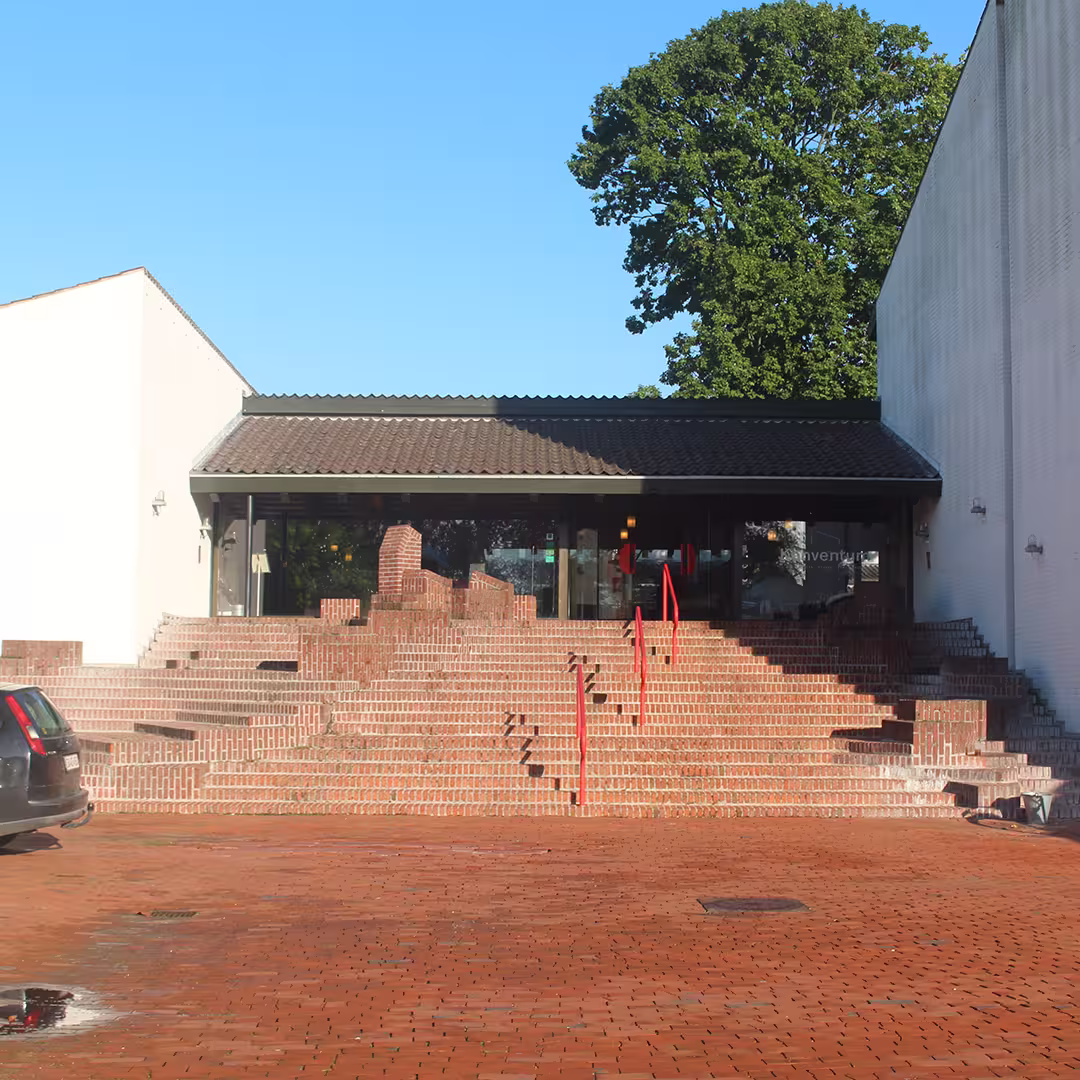 Entrance to the Konventum Convention Centre in Elsinore, Denmark, the venue for Dyalog '19. The entrance features wide brick steps with red handrails leading up to glass doors under a tiled roof, surrounded by white walls and a large tree in the background.