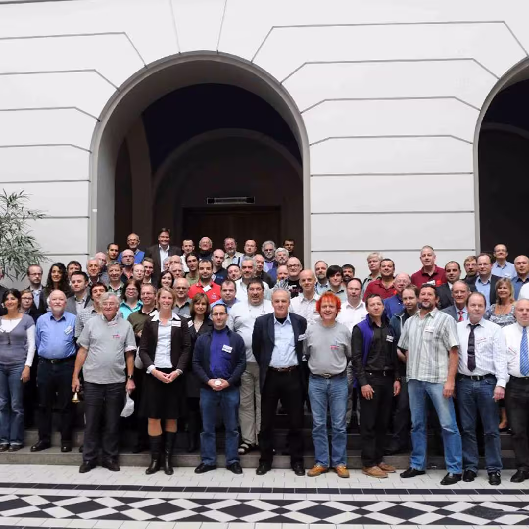A group photo from the Dyalog '10 event in Berlin, Germany, showing a large gathering of delegates posing together in front of an arched entrance within the conference venue. The group includes men and women of various ages, dressed in casual and business attire, many wearing conference badges. The front row stands on the tiled floor, while others stand on steps behind them. The background features white walls, arched doorways, and a potted plant on the left. Everyone is looking at the camera.
