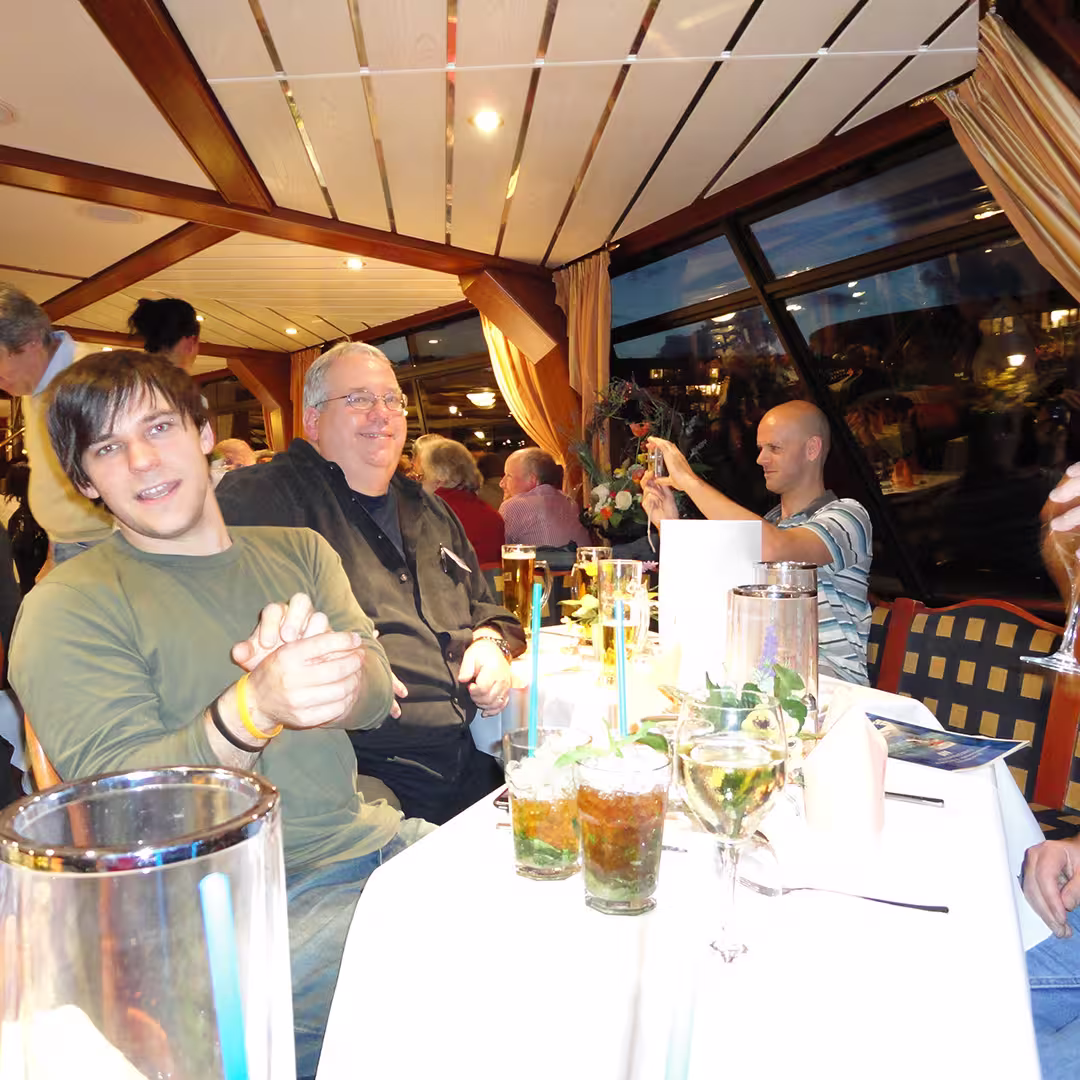 An evening social gathering during the Dyalog '10 event in Berlin, Germany. Three men are seated at a table with drinks, including mojitos with mint and tall beers. The man on the left is wearing an olive green long-sleeve shirt, leaning forward with his hands clasped. The man in the middle, wearing a black shirt and glasses, is smiling while seated back. On the right, Jay Foad, wearing a striped polo shirt, is holding a phone and taking a picture. The table is decorated with flowers and white napkins. The background shows other delegates mingling, with curtains drawn back to reveal windows reflecting the interior lights.