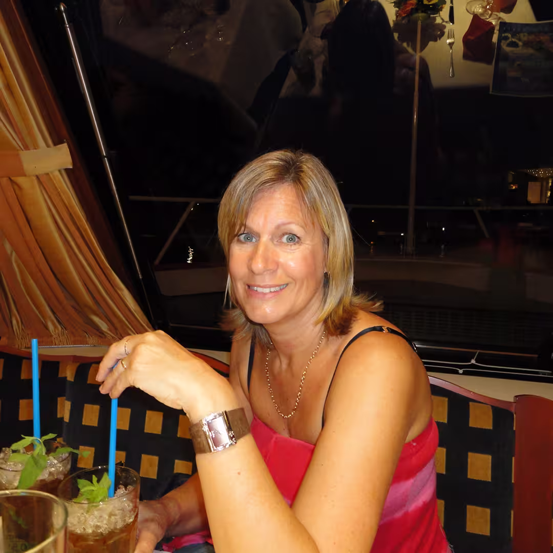 Karen Shaw at an evening social gathering during the Dyalog '10 event in Berlin, Germany. She is seated on a cushioned bench with a geometric pattern, holding a drink garnished with mint and a blue straw. She is wearing a pink sleeveless dress, a silver watch, and a gold necklace, smiling warmly at the camera. The background features a window reflecting the interior lights and curtains drawn to the side.