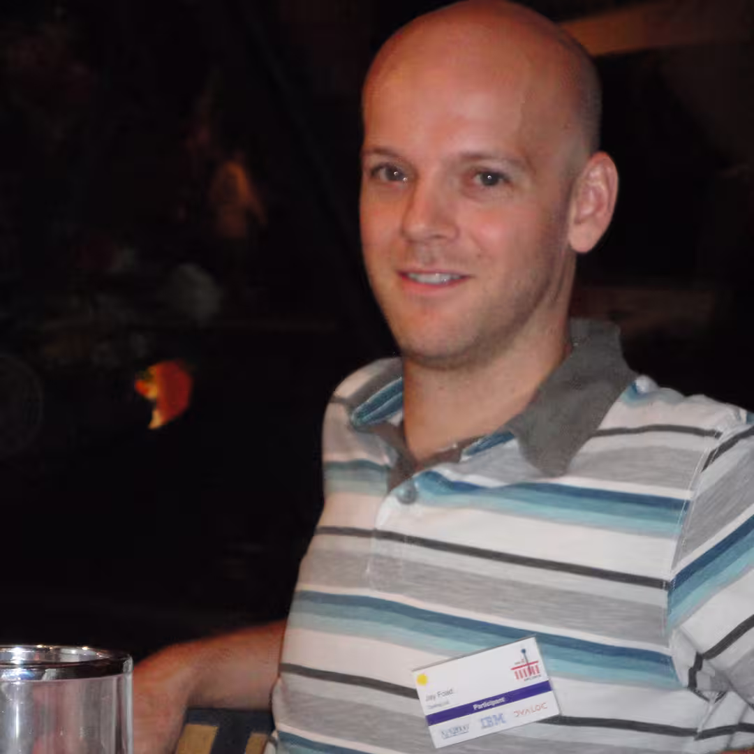Jay Foad, a participant at the Dyalog '10 event in Berlin, Germany, seated at a table during an evening social gathering. He is wearing a striped polo shirt with shades of blue, white, and grey. His conference badge, showing his name alongside logos for Dyalog, IBM, and Vector, is pinned to his shirt. A glass is visible in the foreground, and the background is dimly lit with foliage.