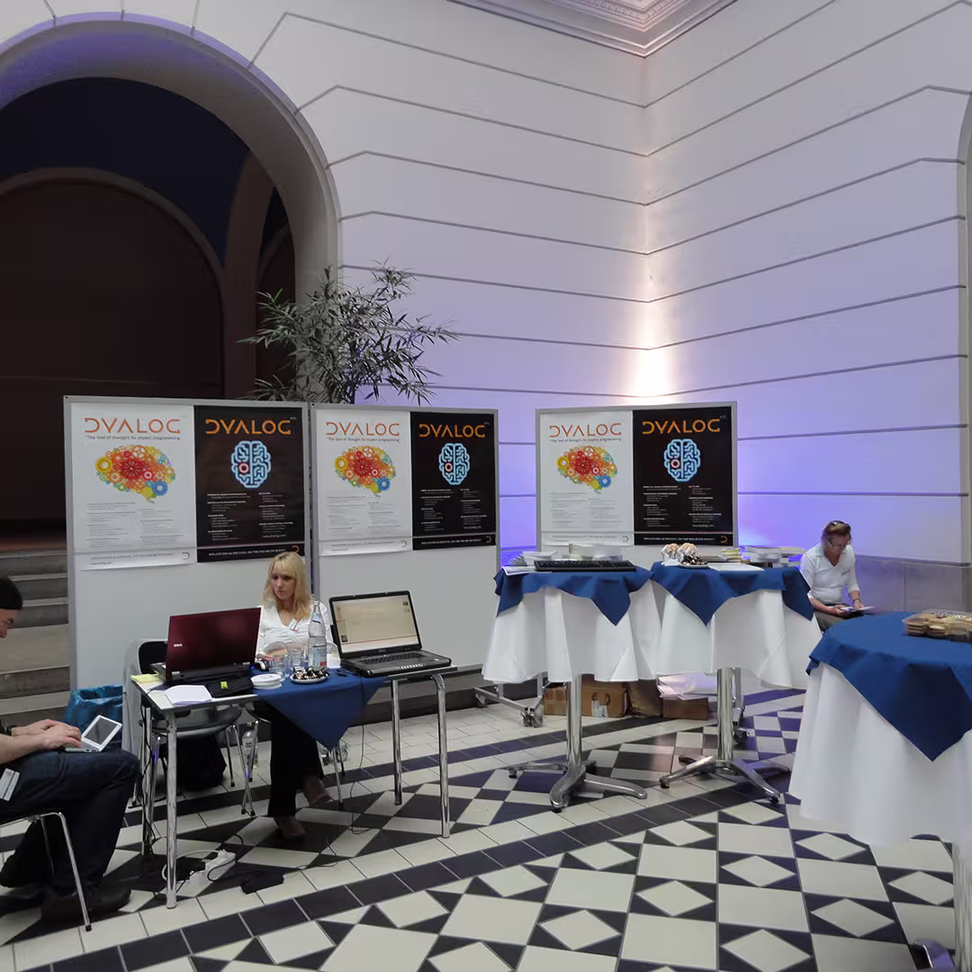 The reception area at the Dyalog '10 event in Berlin, Germany, showcasing Dyalog branding. Five large posters with the "Dyalog" logo and a colourful brain graphic are displayed on partition walls. In front of the booth, a woman with blonde hair sits at a table with two laptops, a bottle of water, and some promotional materials. To the left, a person is seated, using a tablet. To the right, a woman in a white shirt sits at another table with refreshments. The tables are covered with white tablecloths and blue overlays. The background features white walls, an arched doorway, and a potted plant.