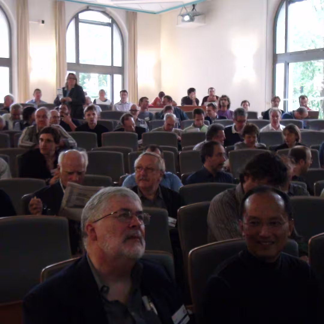 A presentation session at the Dyalog '10 event in Berlin, Germany, showing a lecture hall filled with delegates seated in grey chairs. In the foreground, Roger Hui, wearing a black turtleneck and glasses, is smiling while seated next to another delegate with white hair and a beard, who is attentively looking forward. The middle and back rows are occupied by delegates engaged in quiet conversation, reading materials, or looking towards the front. The room has large arched windows allowing natural light to enter.