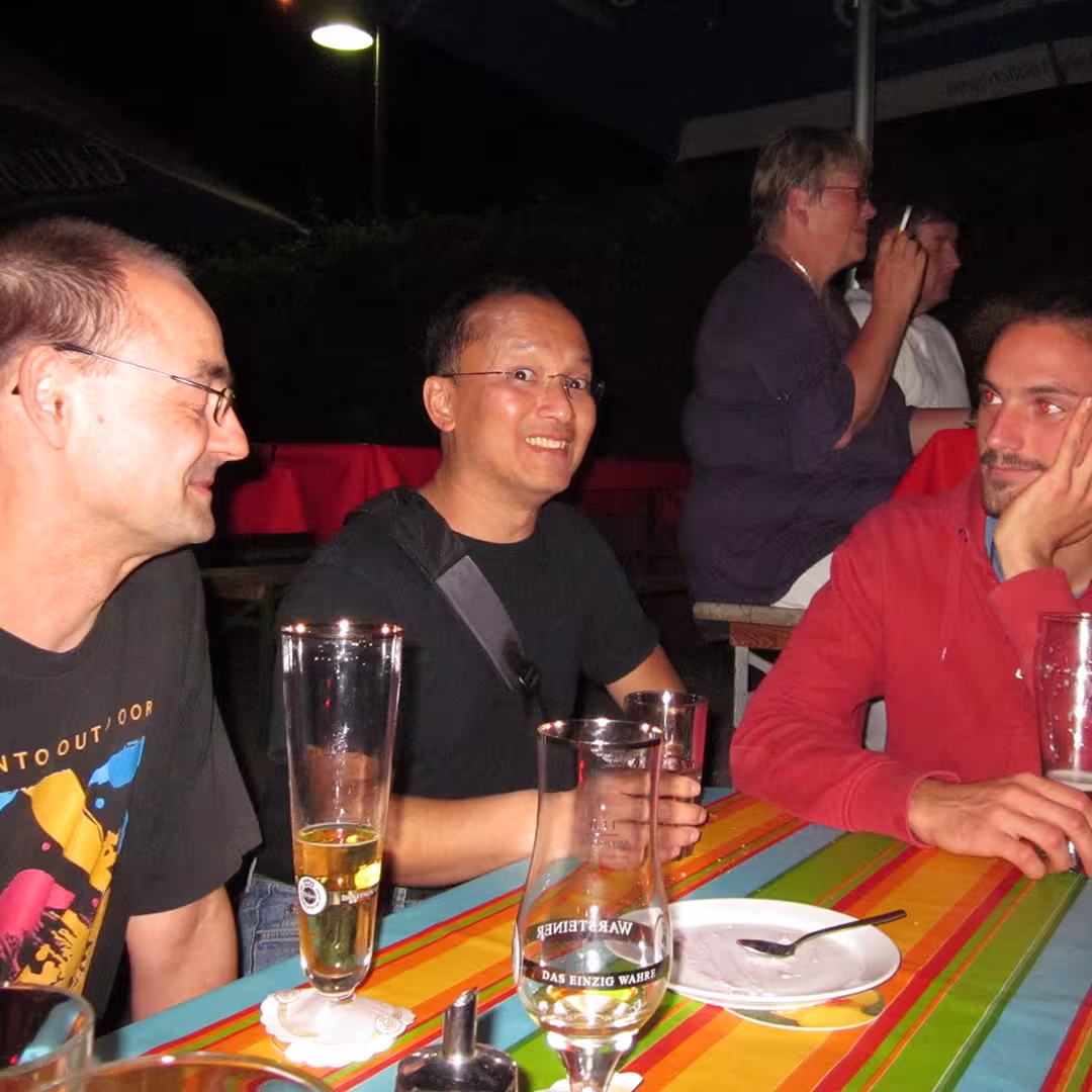 An evening social gathering during the Dyalog '10 event in Berlin, Germany. Three men are seated at a table covered with a colourful striped tablecloth, with tall beer glasses and a dessert plate in front of them. Roger Hui, a prominent figure in the APL community, is in the middle, smiling broadly while holding a glass. To his left, a man in a black t-shirt with a colourful design is turned toward him, smiling. On Roger's right, a man in a red hoodie rests his head on his hand, appearing deep in thought. The setting is outdoors, under dim lighting.