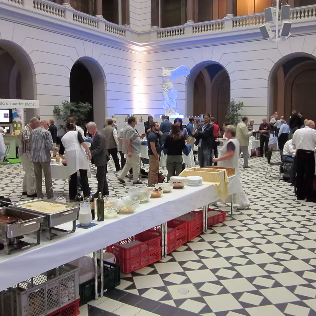 A scene from the Dyalog '10 event in Berlin, Germany, showing delegates gathered in a spacious hall with black-and-white geometric tiled flooring. In the foreground, a long buffet table covered with a white tablecloth holds serving trays, bowls of salads, and bottles of oil and dressing. Red crates are stacked underneath. In the middle ground, groups of delegates stand and chat, while some enjoy refreshments. In the background, a white statue is illuminated with blue lighting, standing against the white walls and archways of the venue.