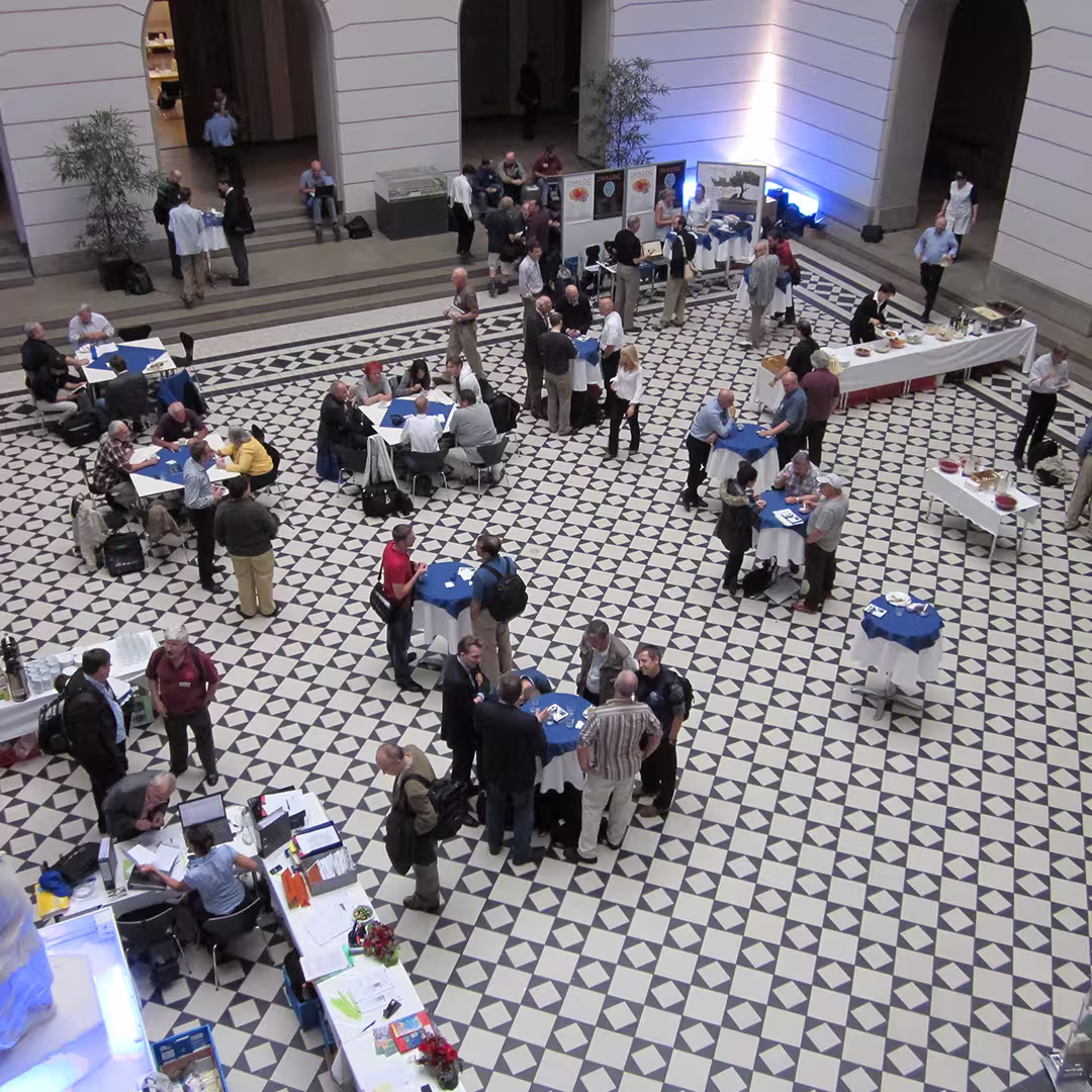 An overhead view of the Dyalog '10 event held in Berlin, Germany, showing a spacious hall with a black-and-white geometric tiled floor. Several groups of delegates are gathered around round tables covered with blue tablecloths, engaged in discussions, working on laptops, or having refreshments. There are exhibition boards and catering tables along the walls, with some delegates standing nearby. The background features tall, white archways and potted plants.