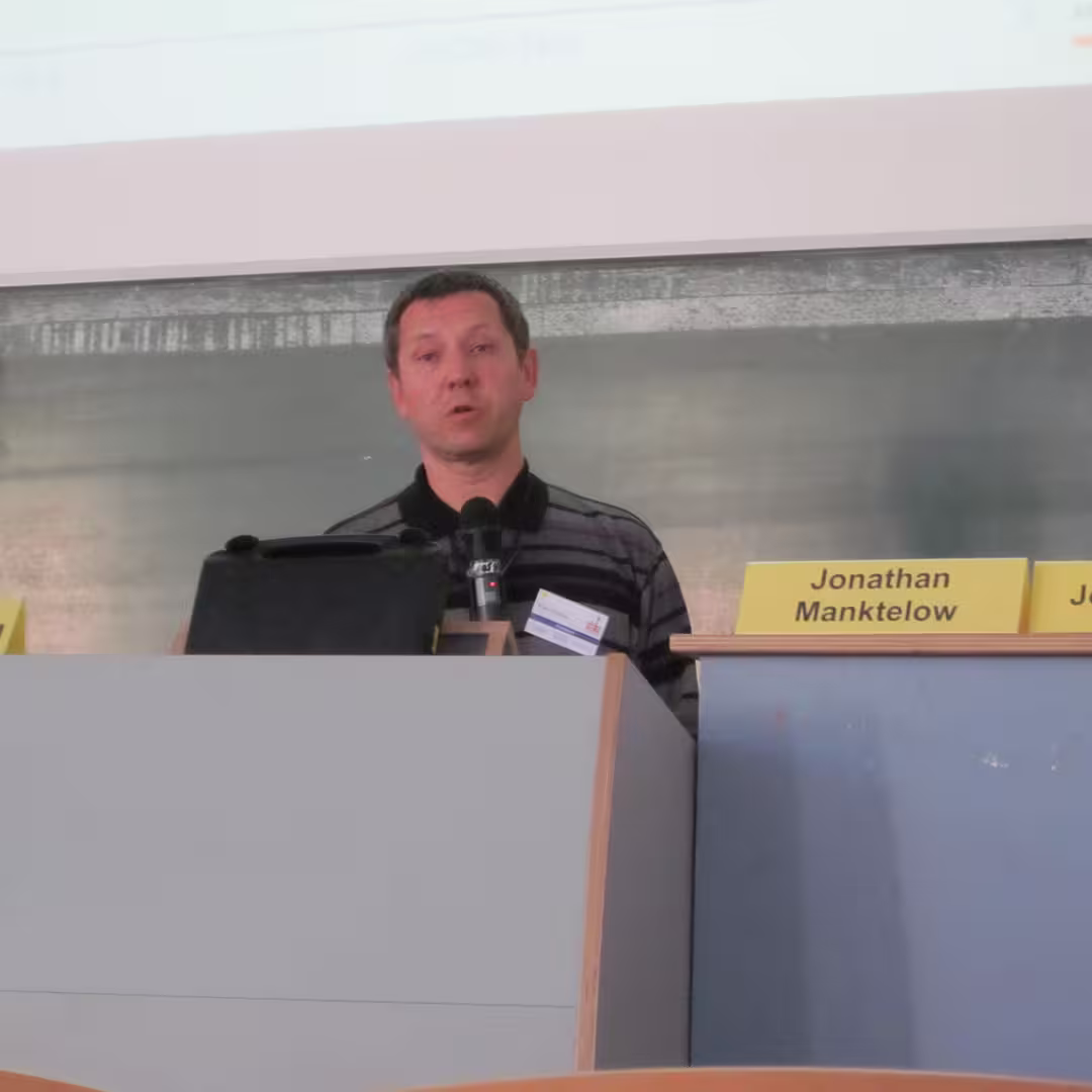 Morten Kromberg presenting at Dyalog '10. He stands at a podium wearing a striped shirt with a microphone attached. A projector screen is visible in the background.
