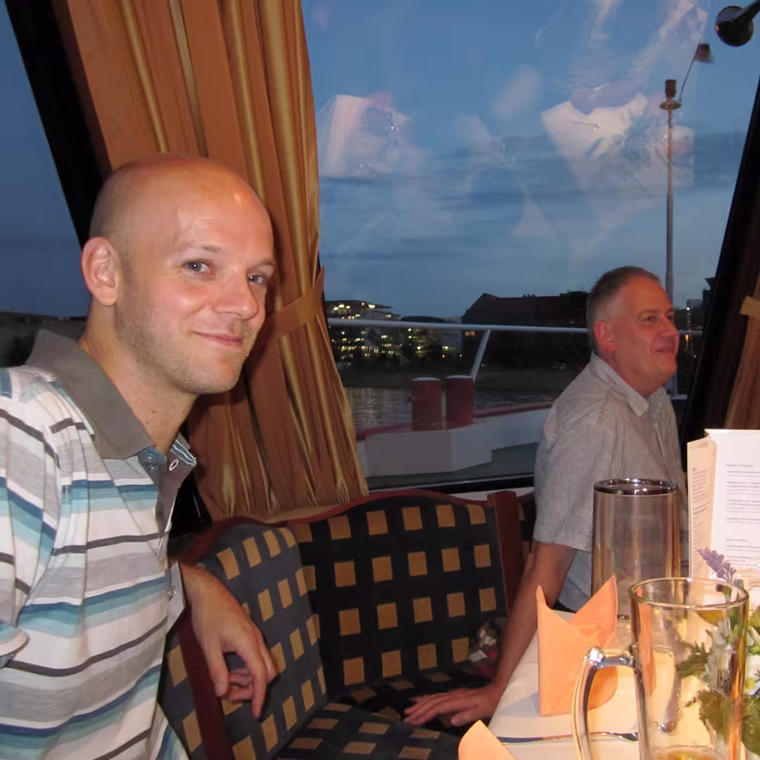 An evening social gathering during the Dyalog '10 event in Berlin, Germany. Jay Foad, wearing a striped polo shirt, sits on the left side of the image, smiling at the camera. Behind him, Andy Shires, wearing a light grey short-sleeve shirt, is seated at the same table, looking off to the side. The table is set with folded orange napkins, a glass jug, and a floral centerpiece. Through the large window behind them, a river, buildings with illuminated windows, and the evening sky are visible. The curtains are drawn to the side, and reflections of the interior lights can be seen on the window.