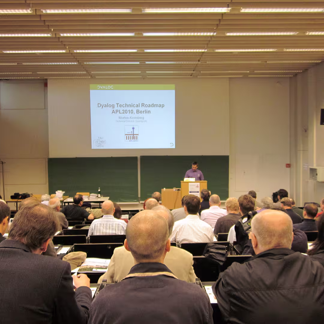 Morten Kromberg presenting the "Dyalog Technical Roadmap" at the Dyalog '10 event in Berlin, Germany. He stands behind a podium in a lecture hall, speaking to an audience of delegates seated in rows. The presentation slide projected on the screen reads "Dyalog Technical Roadmap, APL2010, Berlin" with Morten Kromberg's name and title as Technical Director of Dyalog Ltd. The room has a large green chalkboard behind the screen and a ceiling with fluorescent lights. Some attendees are taking notes while others are listening.