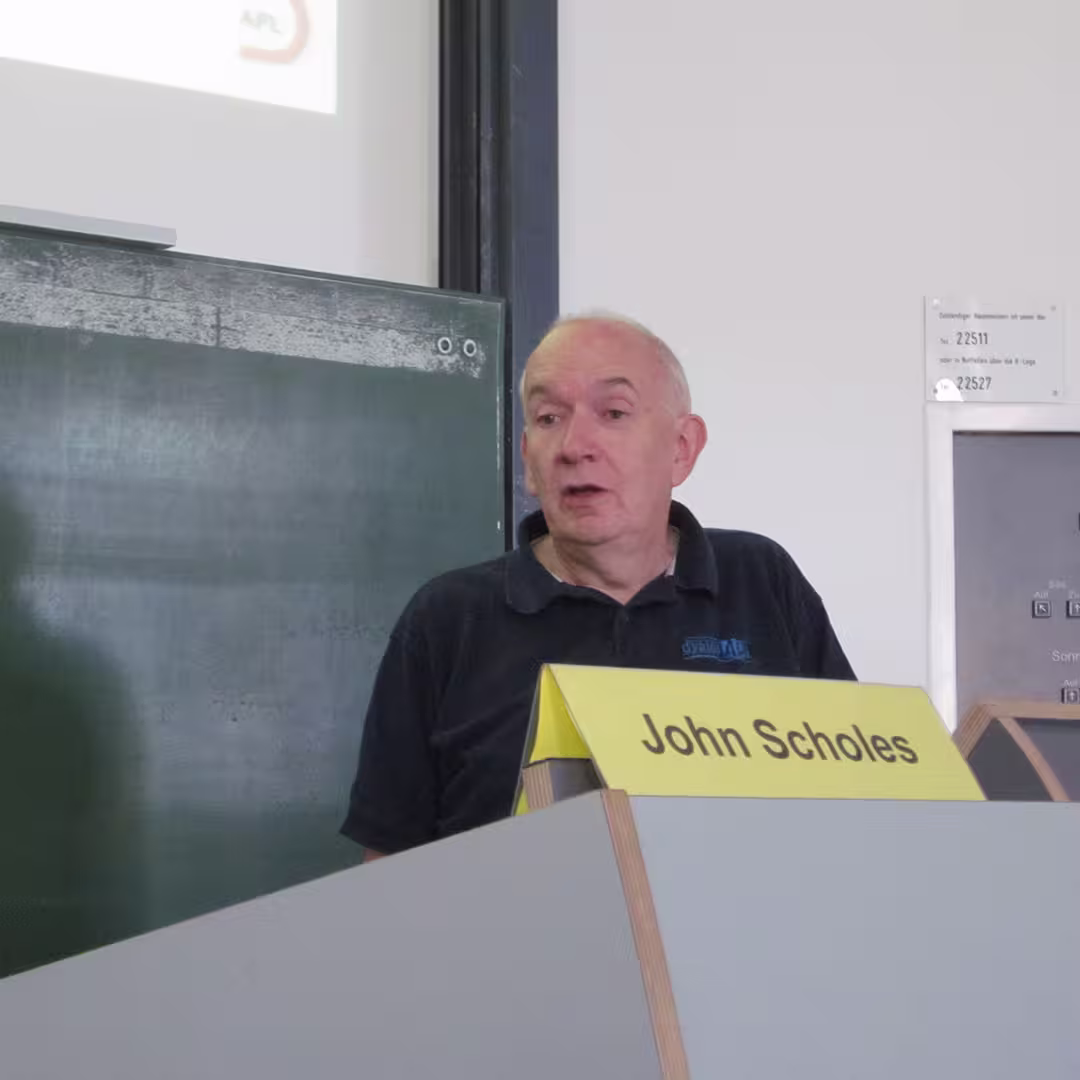 John Scholes giving a presentation at the Dyalog '10 event in Berlin, Germany. He is standing behind a podium with a yellow nameplate that reads "John Scholes." He is wearing a dark blue polo shirt with the Dyalog logo embroidered on the left side. Behind him is a large green chalkboard with faint chalk marks, and a projector screen displaying a partial logo in the top left corner. The background features a white wall with signage and control panels.
