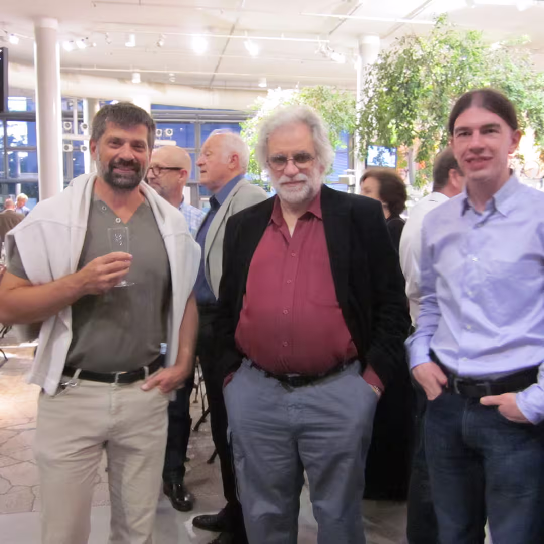 Geoff Streeter, Bjørn Christensen, and another attendee at the Dyalog '10 event in Berlin, Germany, standing together during an evening social gathering. Geoff Streeter is in the middle, wearing a burgundy shirt, grey trousers, and a black jacket, with his hands in his pockets. Bjørn Christensen is on the left, wearing a grey t-shirt with a light grey jumper draped over his shoulders, holding a champagne flute and smiling. The delegate on the right is wearing a light purple shirt and dark jeans. The background shows other attendees mingling, with large potted plants and bright overhead lights in the modern venue.