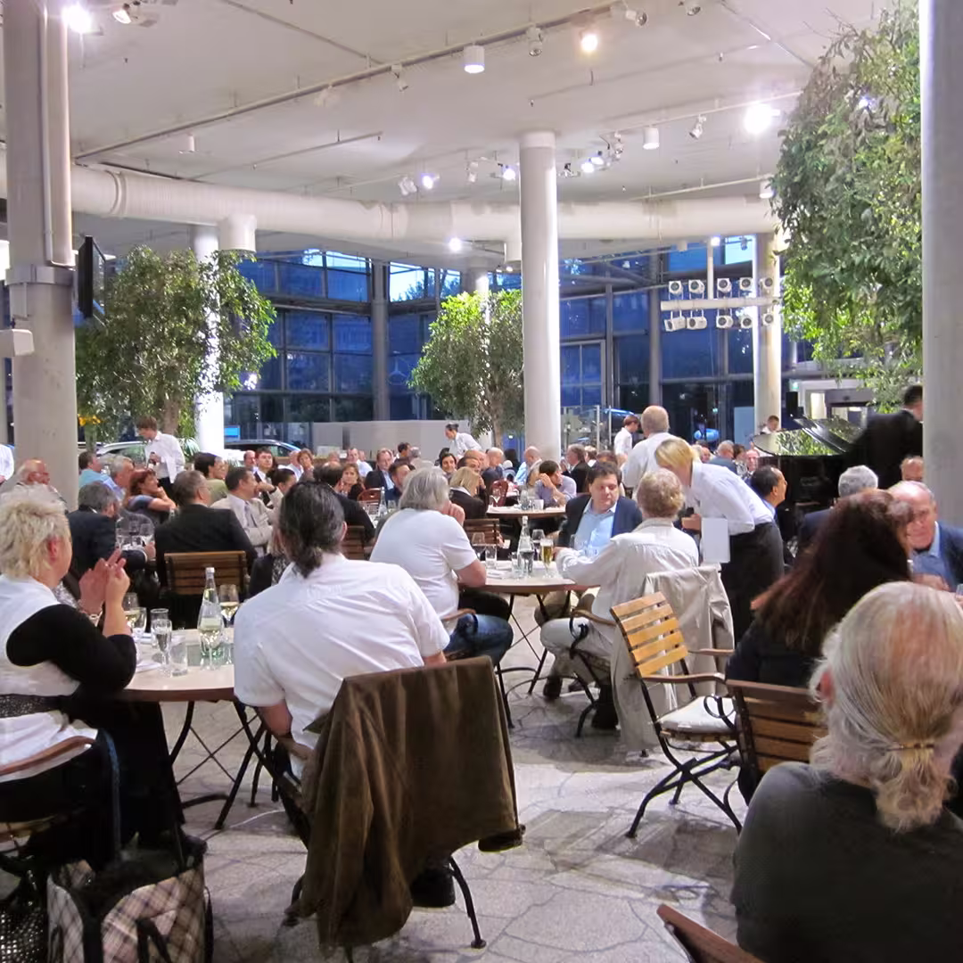 A large dinner gathering during the Dyalog '10 event in Berlin, Germany. Delegates are seated at multiple round tables in a spacious, modern venue with tall windows, concrete pillars, and large potted plants. The tables are set with wine glasses, water bottles, and plates. In the foreground, a woman with short blonde hair sits at a table, clapping, while a man with long dark hair, wearing a white shirt, sits nearby with a jacket draped over the back of his chair. Waitstaff are seen attending to guests, and a grand piano is visible on the right side of the image. The background shows more attendees engaged in conversation under bright overhead lighting.