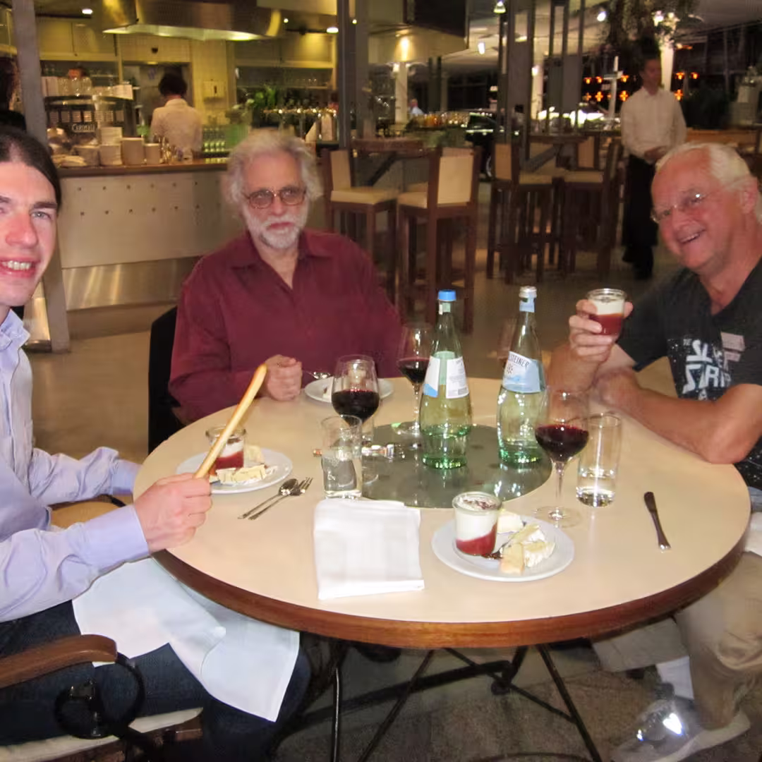 A dinner gathering during the Dyalog '10 event in Berlin, Germany. Three men are seated around a table enjoying dessert and drinks. Geoff Streeter is in the middle, wearing a burgundy shirt and glasses, with a glass of red wine in front of him. Chris Lincoln is on the right, wearing a black t-shirt with a graphic design, holding a dessert glass while smiling. On the left, a man in a light purple shirt is holding a breadstick and smiling at the camera. The table is set with plates of dessert, wine glasses, water bottles, and cutlery. The background shows a modern dining area with a bar, high chairs, and other diners.