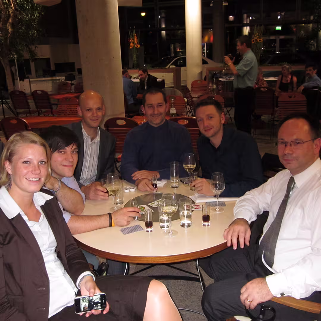 A group of six people seated around a table during the Dyalog '10 event in Berlin, Germany, engaged in conversation and enjoying drinks. Richard Smith of Dyalog is on the far right, wearing glasses, a white shirt, and a tie, smiling at the camera. The table is set with wine glasses, water glasses, and small bottles. A woman in a brown blazer and white shirt sits in the foreground, holding a smartphone. Next to her, a man in a light blue shirt with dark hair leans in, smiling. Jay Foad, wearing a grey pinstriped suit, is seated next to a man in a blue fleece jacket. Another man in a dark shirt sits beside Richard. The background shows other tables, large concrete pillars, and windows reflecting the interior lights. Dan Baronet is lurking in the background.