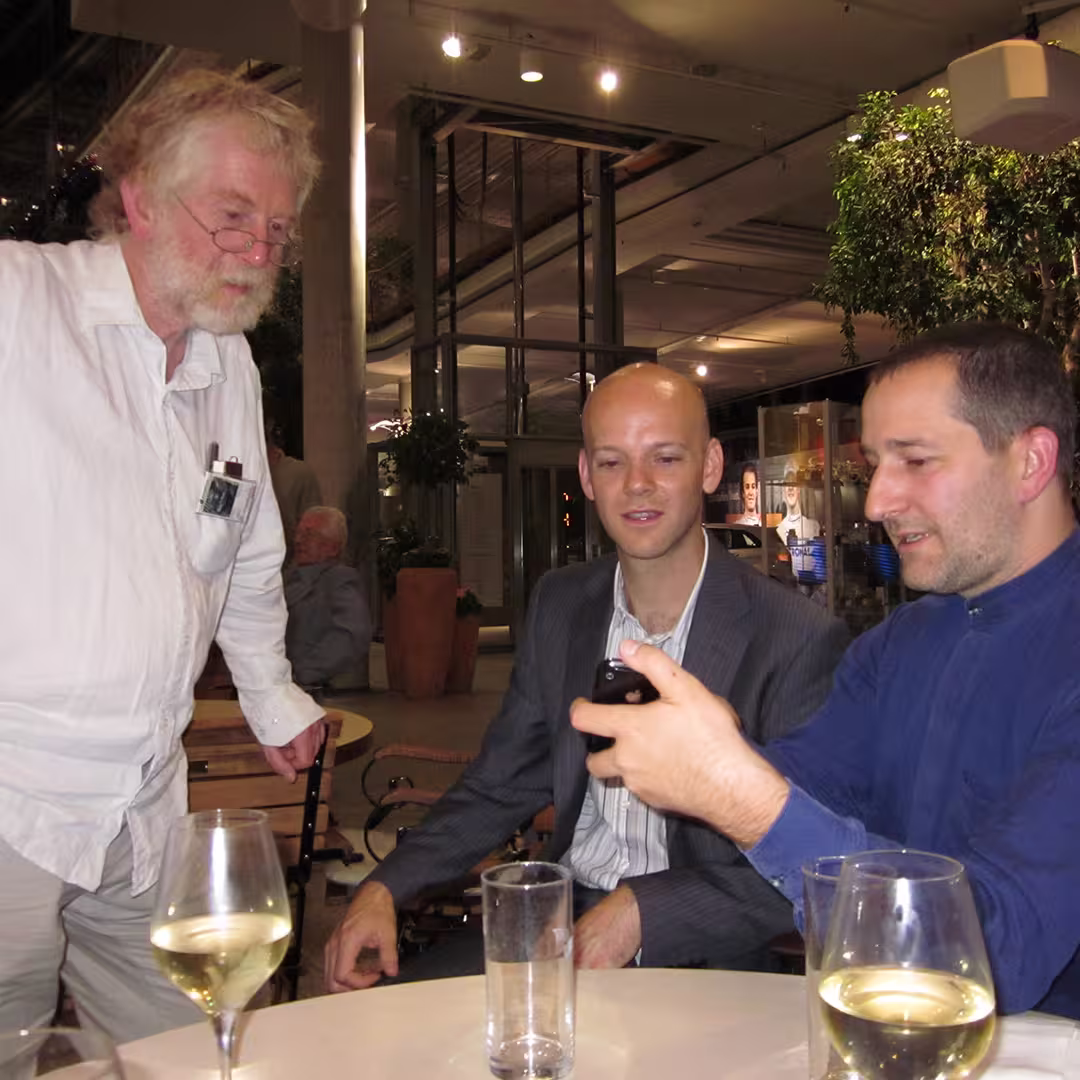 An evening social moment during the Dyalog '10 event in Berlin, Germany. Three men are gathered around a table with wine glasses and water. On the left, an older man with white hair, a beard, glasses, and a white shirt stands while leaning slightly forward. In the middle, Jay Foad, wearing a grey pinstriped suit, is seated, looking at a smartphone held by the man on the right, who is dressed in a blue fleece jacket. The setting is indoors, with large potted plants, glass windows, and bright overhead lighting in the background.