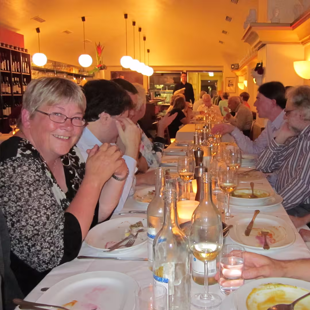 Gitte Christensen of Dyalog, smiling while seated at a long dining table during an evening dinner at the Dyalog '10 event in Berlin, Germany. She is wearing glasses and a black and white floral top. The table is filled with plates, wine glasses, water bottles, and candles. Other delegates are seated along the table, engaged in conversation. The restaurant has warm lighting, with spherical pendant lights hanging from the ceiling and shelves of wine bottles lining the walls. In the background, a waiter stands near the kitchen, and more diners are visible at the far end of the table.
