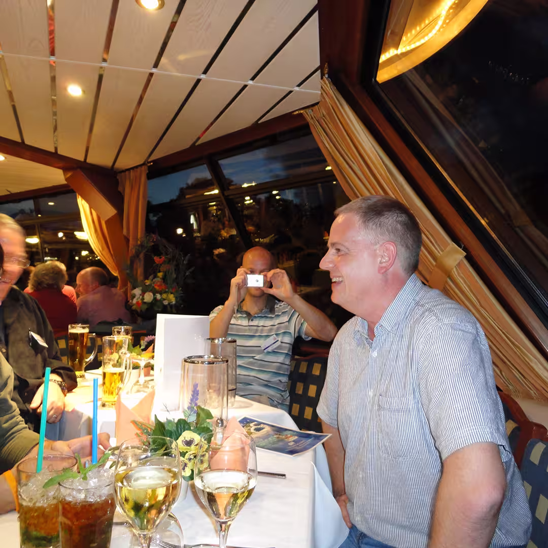 Andy Shires at an evening social gathering during the Dyalog '10 event in Berlin, Germany. He is seated at a table, smiling and engaged in conversation, wearing a striped short-sleeve shirt. In the background, Jay Foad, wearing a striped polo shirt, is taking a photo with a small camera. The table is set with glasses of wine, mojitos with mint and blue straws, a floral centerpiece, and a menu. The setting appears to be on a boat or near a large window, with reflections of lights visible outside. The ceiling has wooden beams, and the curtains are drawn back.