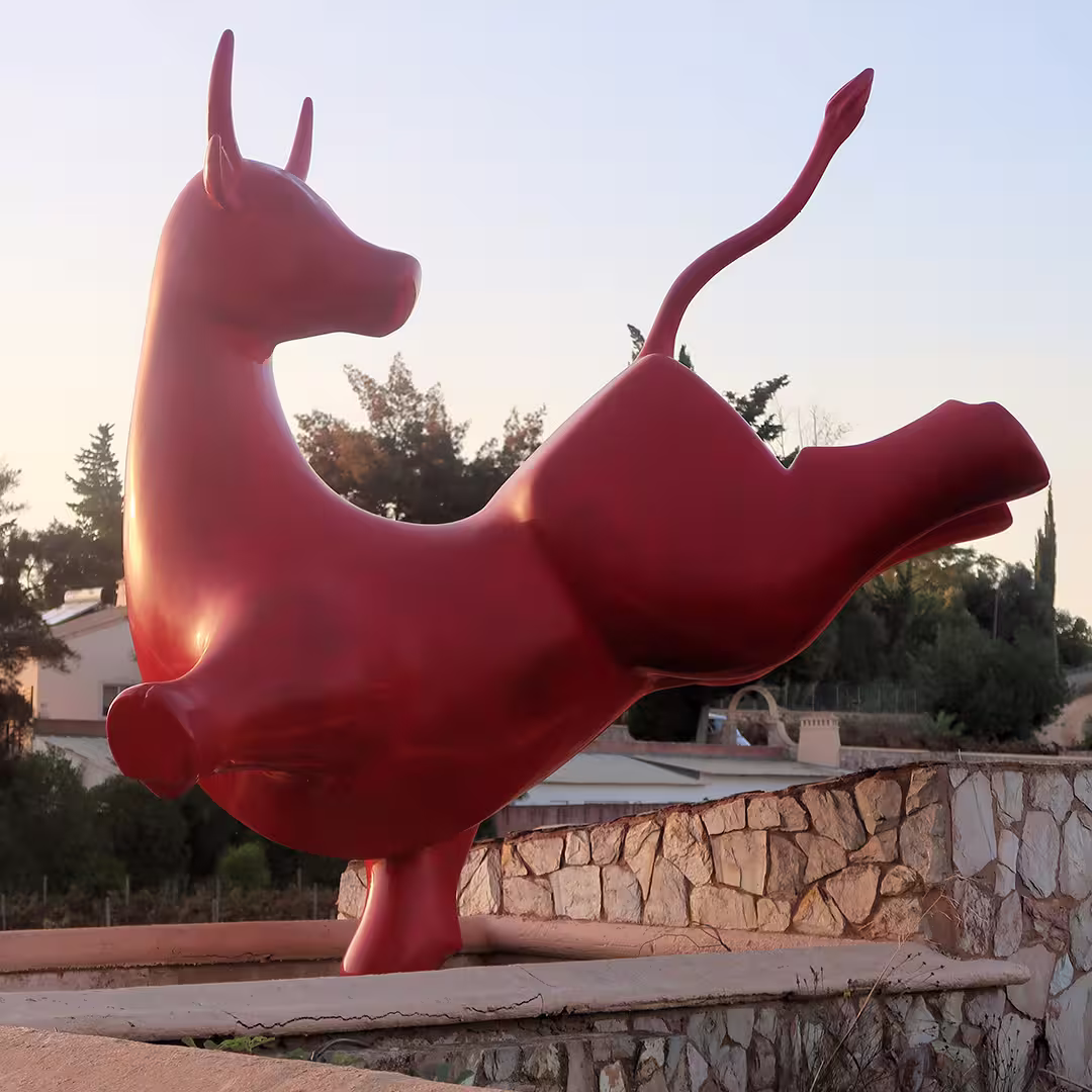 A large, modern red bull sculpture stands on one hoof atop a stone pedestal. The bull has a sleek, stylised design with smooth curves and an elongated tail that arches upward. The background features lush greenery, trees, and a few buildings, suggesting an outdoor setting in Olhão, Portugal, during the Dyalog '22 event. The sky is clear.
