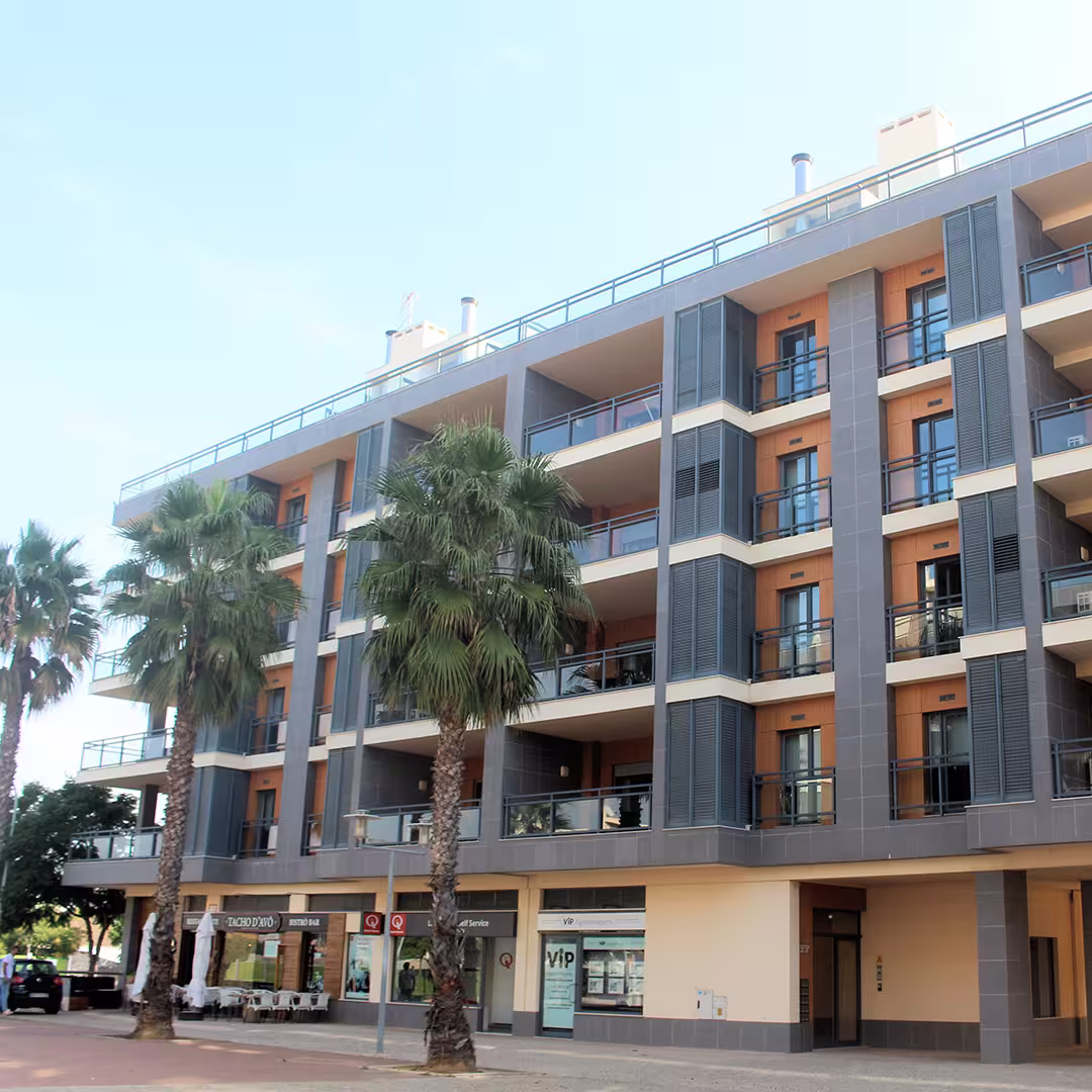 The Real Marina Hotel & Spa in Olhão, Portugal, features a modern, multi-storey facade with balconies and large windows. The building's exterior combines grey panels with warm wooden accents, and tall palm trees line the front. The ground floor hosts a few shops and a café with outdoor seating. The sky above is clear and bright.