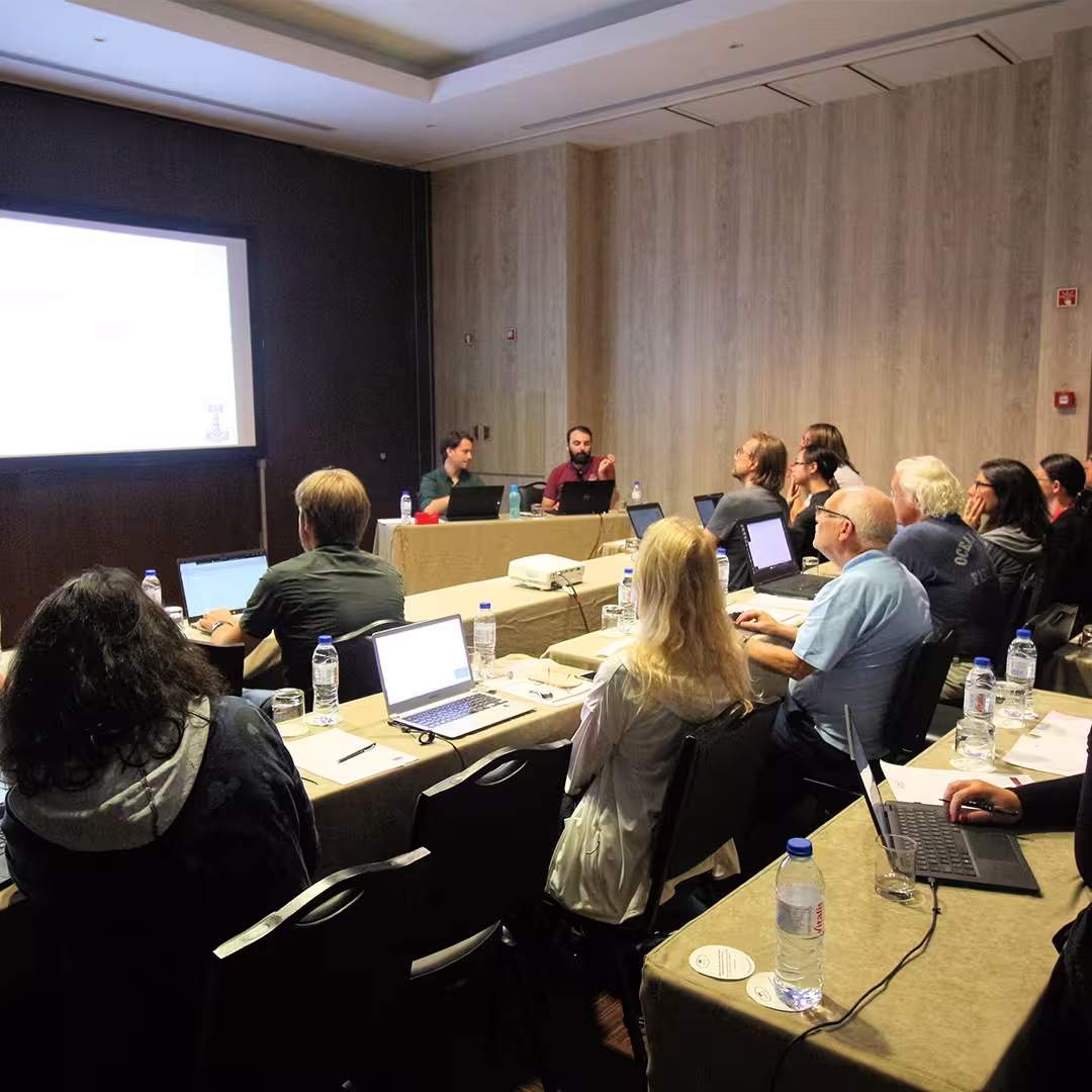 Delegates at Dyalog '22 sit around tables in a conference room, participating in a workshop. Laptops, notebooks, and water bottles are scattered across the tables. At the front, Rodrigo Girão Serrão (on the left) listens while Rich Park (on the right) speaks, with a projection screen displaying content behind them. The room has wood-panelled walls and soft lighting, creating a focused yet comfortable atmosphere.