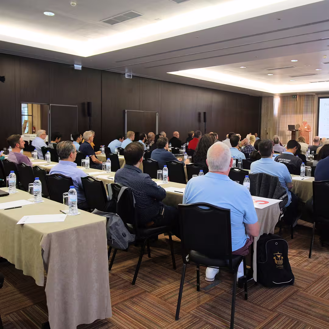 Delegates at Dyalog '22 sit in rows of tables, attentively listening to Gitte Christensen presenting at the front of the conference room. Each table is set with water bottles, notepads, and pens. The room has dark wood-panelled walls, a carpeted floor, and soft ceiling lighting. A large screen beside the podium displays the presentation slides.