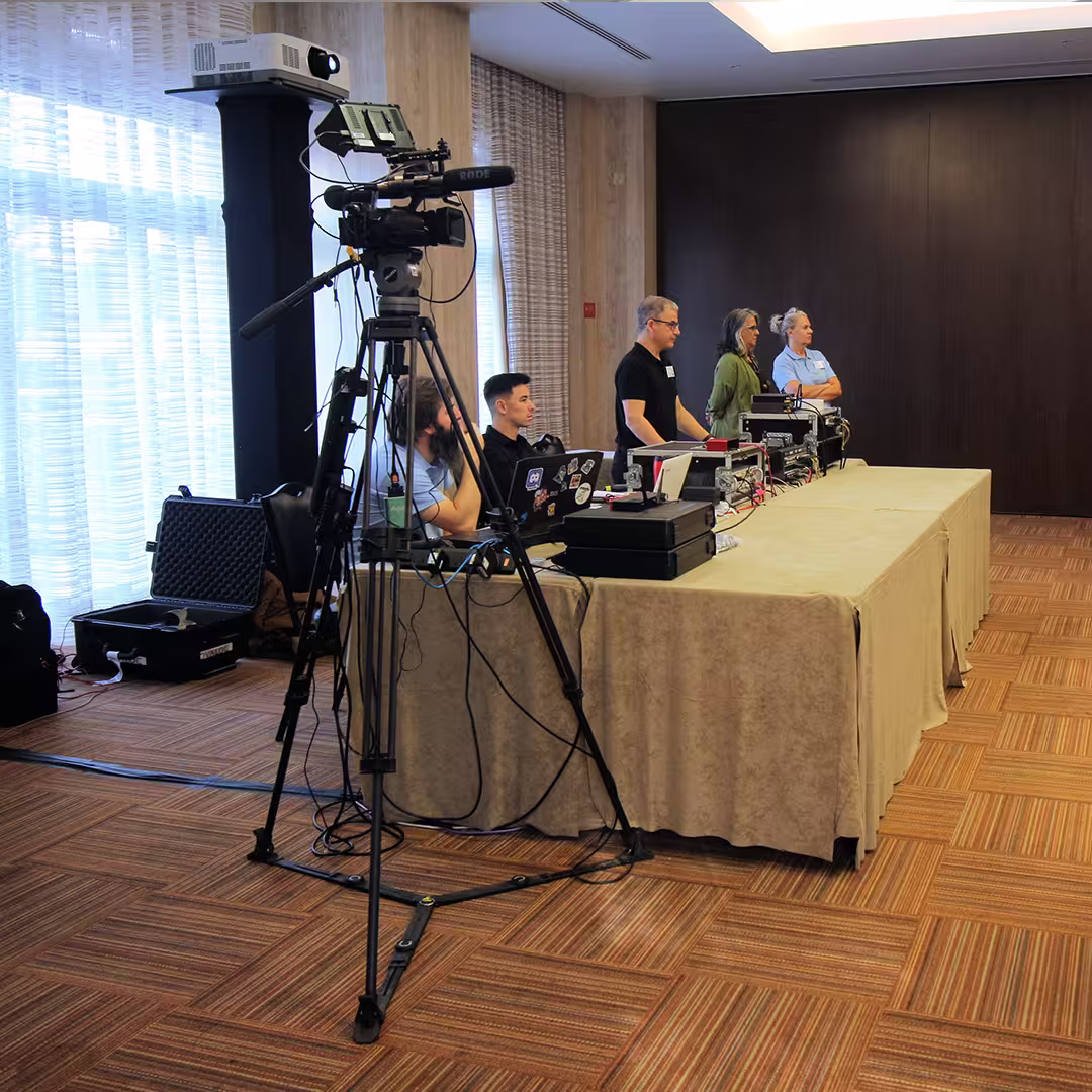 A camera crew operates the AV setup at Dyalog '22. A professional video camera on a tripod stands in the foreground, aimed at the conference stage. Behind it, two people sit at a table covered with laptops, cables, and other technical equipment, while three others stand nearby, monitoring the setup. Large windows with sheer curtains allow natural light into the room, which has wood-panelled walls and a patterned carpet.