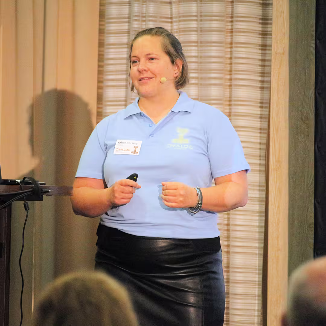 Stine Kromberg, CEO of Dyalog Ltd, stands on stage at Dyalog '22, delivering a presentation. She wears a light blue Dyalog polo shirt and a black leather skirt, holding a presentation remote in her hand. A headset microphone is positioned near her mouth. Behind her, beige curtains and a wood-panelled wall form the backdrop. The edge of an audience member's head is visible in the foreground.